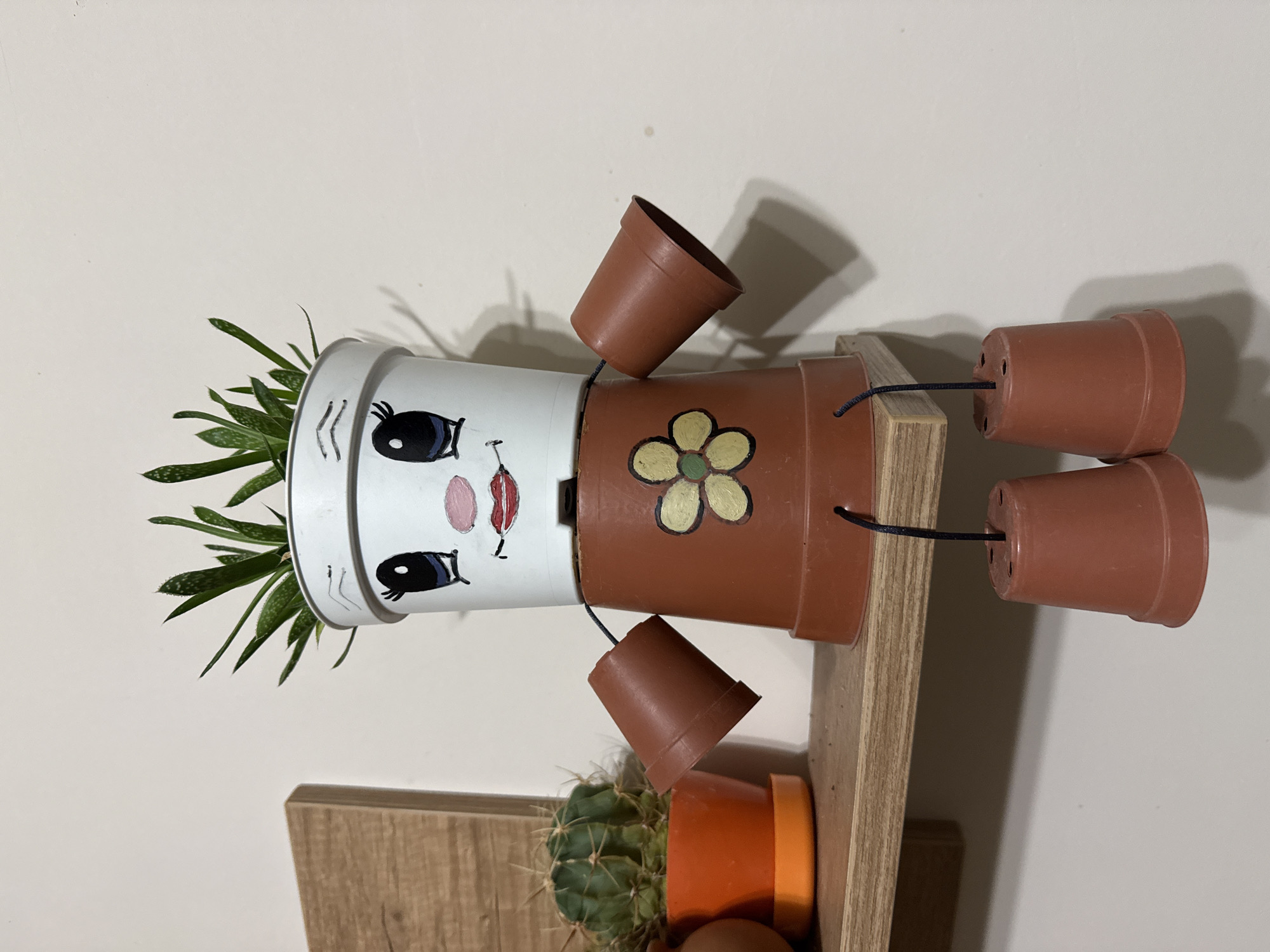 A decorative figure, crafted from repurposed plant pots, is situated on a light-colored wooden shelf against a plain, light-colored wall. This scene is located in Stara Zagora, Bulgaria. The figure's head is a white plastic pot containing a green, spiky succulent plant, visually styled as hair. A cartoonish face is painted on the white pot, featuring large black eyes with white pupils, pink blush circles on the cheeks, a small pink nose, and red lips. The body is an inverted, larger brown plastic pot, painted with a yellow and green flower design on its front. Two smaller, horizontally oriented brown plastic pots are attached to the upper sides of the body pot by black strings, serving as arms. Two identical small brown plastic pots hang vertically beneath the body, suspended by black strings threaded through holes in the shelf and the pots, forming legs. To the left of the figure, on the same wooden shelf, rests a small, spherical green cactus in a bright orange plastic pot with a matching orange saucer. The wall behind the shelf is unadorned and light beige or off-white. The lighting appears uniform, casting subtle shadows to the right and beneath the figure.