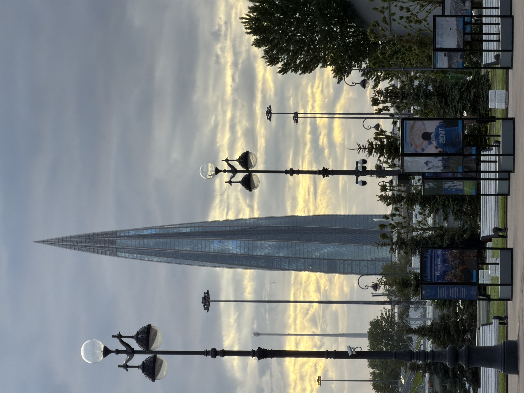 The image captures a scene in Saint Petersburg, Russia, featuring the Lakhta Center skyscraper, a modern glass and steel structure that tapers to a point. The sky is overcast, with streaks of light breaking through the clouds. In the foreground, there are ornate street lamps with spherical lights. Additionally, there are structures with informational panels and benches, likely a public display. The perspective is from a low angle, looking upwards towards the tower and the sky. The environment is urban, with trees and vegetation present alongside the built infrastructure.