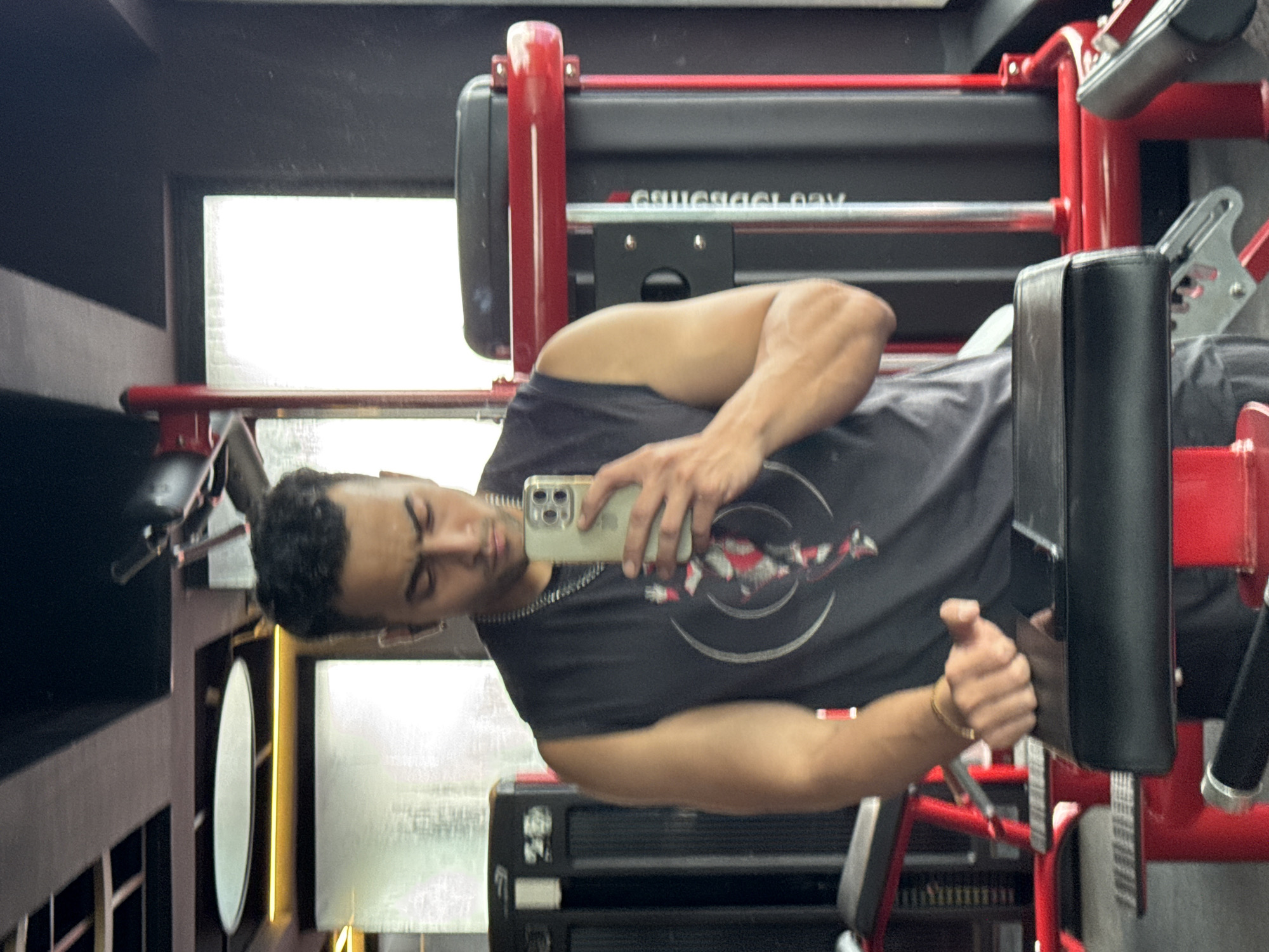 In a gym located in Kolkata, India, a man is taking a selfie. He is wearing a sleeveless black t-shirt and a silver chain. He is holding a smartphone in his right hand, using it to capture his reflection in a mirror. The man's left hand is resting on a piece of gym equipment, possibly a bench. The gym environment is visible in the background, with red-colored exercise machines and black cabinetry. The lighting appears to be a mix of artificial gym lights and natural light coming from a window.