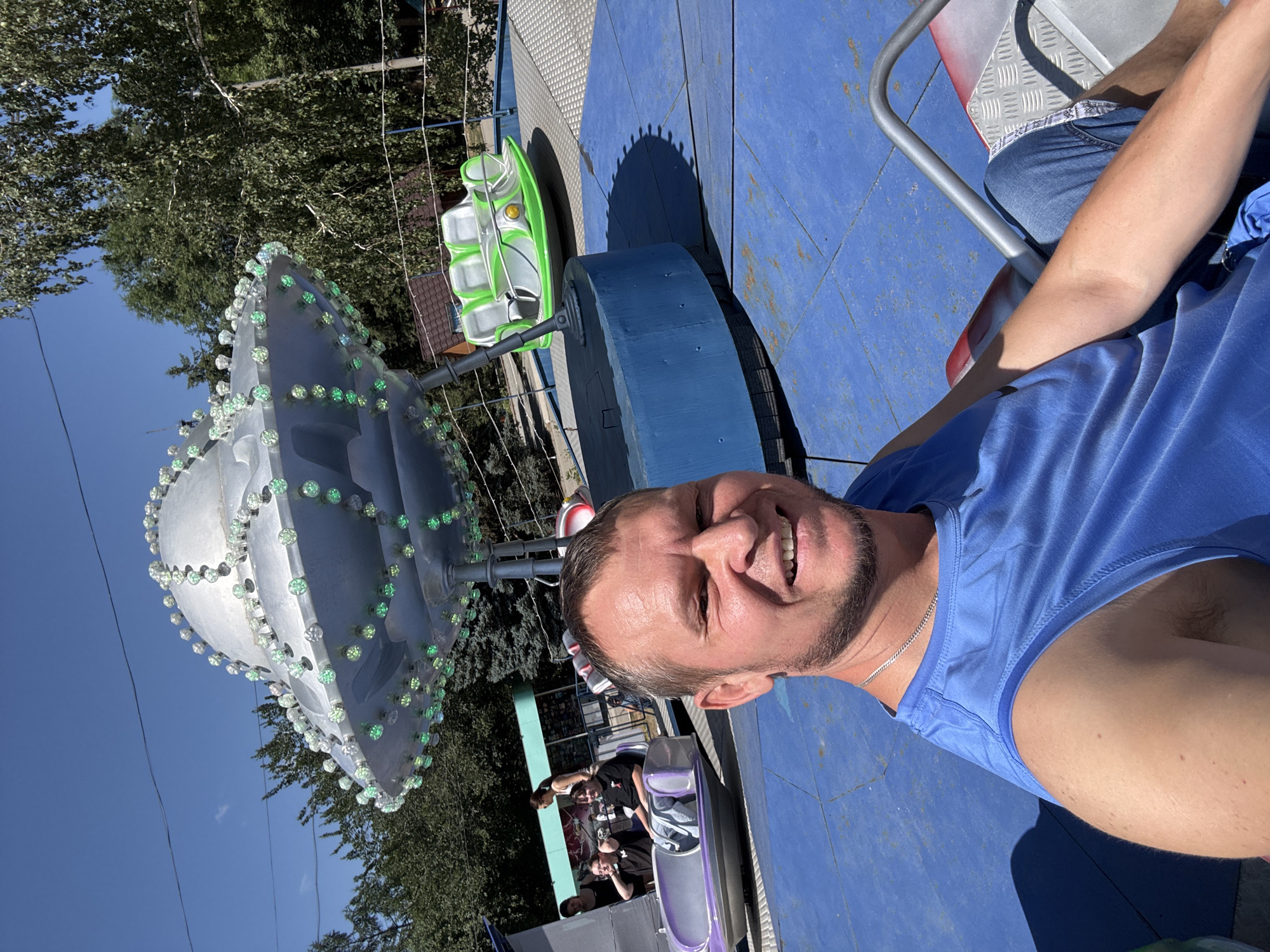In an amusement park setting, a man with short brown hair and stubble is taking a selfie, smiling at the camera. He is wearing a blue sleeveless t-shirt and blue jeans. Behind him, a silver, UFO-shaped amusement park ride is visible, decorated with green and white lights. Several other amusement park rides, including a green and white multi-seated ride and a purple vehicle, are also present. In the background, trees and a building can be seen, under a clear blue sky. The image is captured in Oskemen, Kazakhstan.