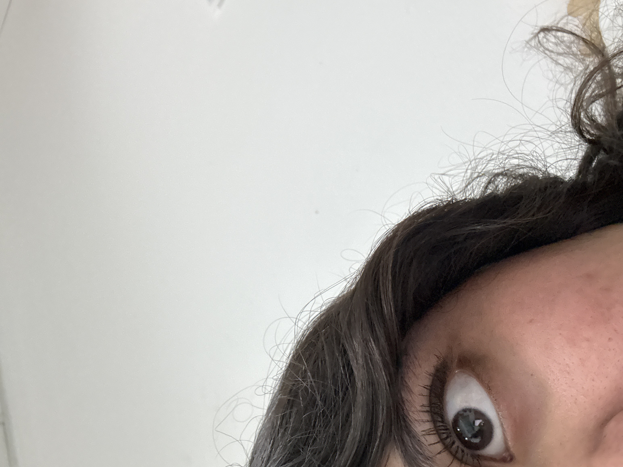 A close-up, asymmetrical view of a person's face is presented against a predominantly white background. The focus is primarily on the right eye, which is wide open and gazing upwards. The skin appears fair and displays freckles, particularly on the forehead. Dark brown hair with loose curls frames the face. No information regarding the city of Puebla, Mexico, is discernible from this image.