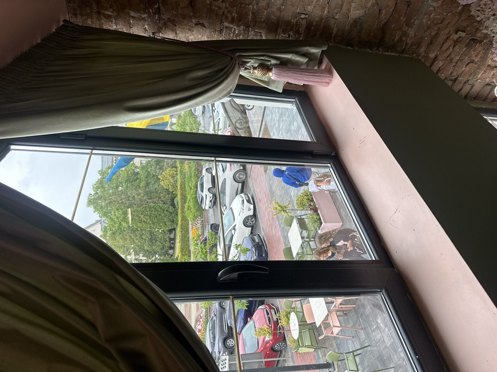 This is a photograph taken from inside a building, looking out through a large window. The window overlooks an outdoor seating area with tables and chairs, and beyond that, a street with parked cars.
On the left side of the image, a green curtain is drawn back, revealing the view. The outdoor seating area has several tables, some with green chairs and some with pink chairs. There are planters with greenery and some flowers.
The street visible through the window has several cars parked along it. These include a red minivan, a dark blue sedan, and several white cars. Trees and buildings are visible in the background. There are two flags visible, one blue and one yellow, suggesting this location is in Ukraine.
In the foreground, near the window, there are two young women standing and looking at a phone. Further to the right, a person wearing a blue hooded jacket is walking away from the camera.
The interior of the building is partially visible, with a pink windowsill and a brick wall on the right side of the frame. A decorative tassel hangs from the curtain. The overall lighting suggests an overcast day. The scene appears to be a cafe or restaurant with outdoor seating.
