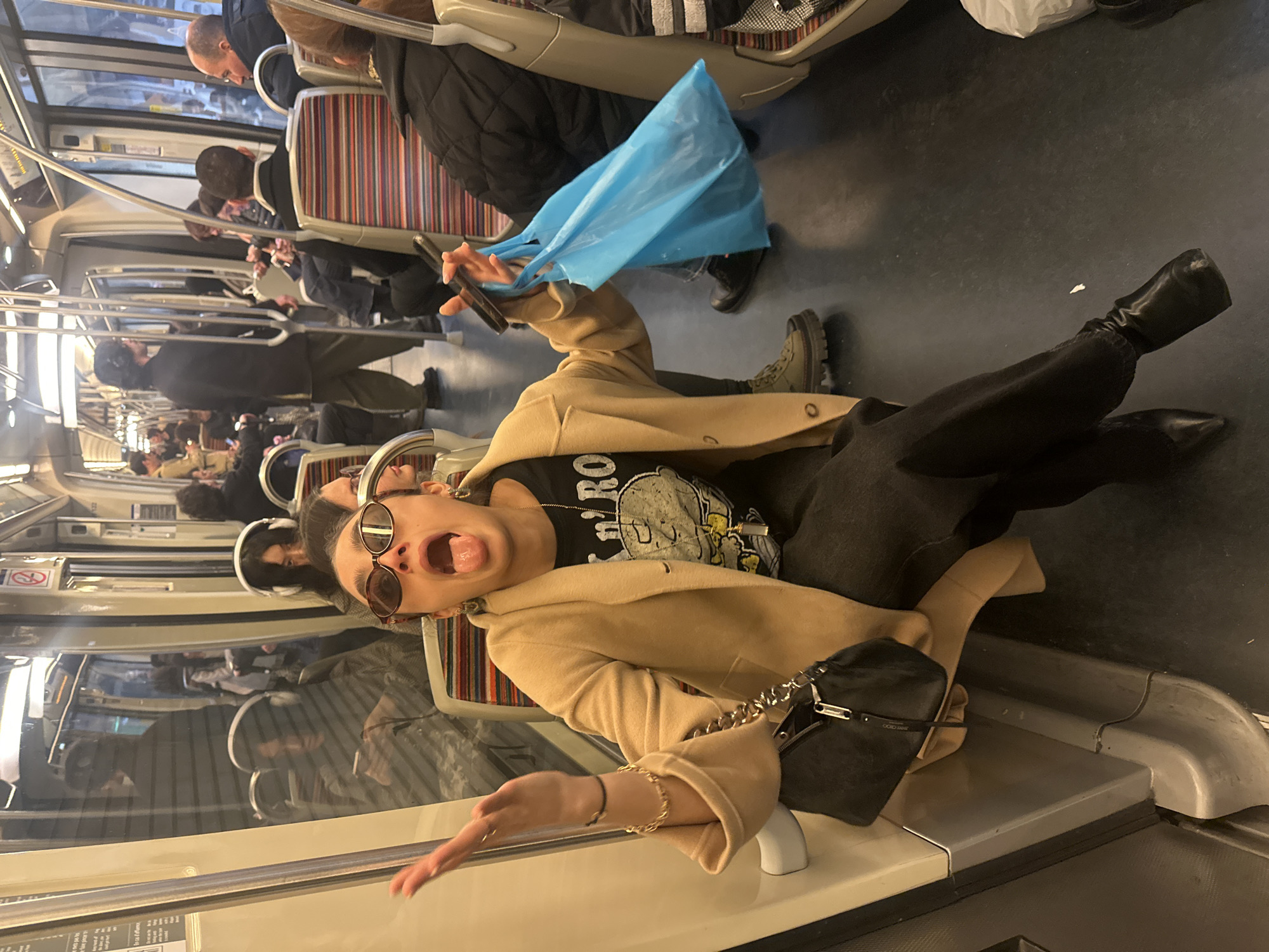 A woman lies across a seat in a train carriage. She is wearing black pants, black boots, and a tan overcoat, with a black purse lying on the seat. She is wearing sunglasses and is holding a light blue plastic bag in one hand. Her mouth is open and her tongue is sticking out. The train carriage is full of passengers. This scene could be occurring in the city of Paris, France.