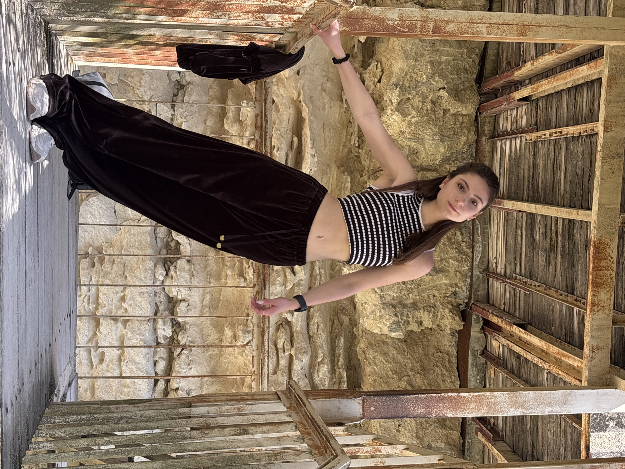 A young woman in a black and white striped top and black velvet pants is hanging from a rusty metal structure within a stone and wooden tower in Varna, Bulgaria. She is holding onto the top rail of the structure with one hand, with a black hair tie on her wrist. Her other arm is extended downward, adorned with a smartwatch. The tower's walls are constructed of weathered stone, while the interior frame is made of aging wood and rusty metal. The perspective suggests the photo was taken from below, looking upward towards the tower's opening.