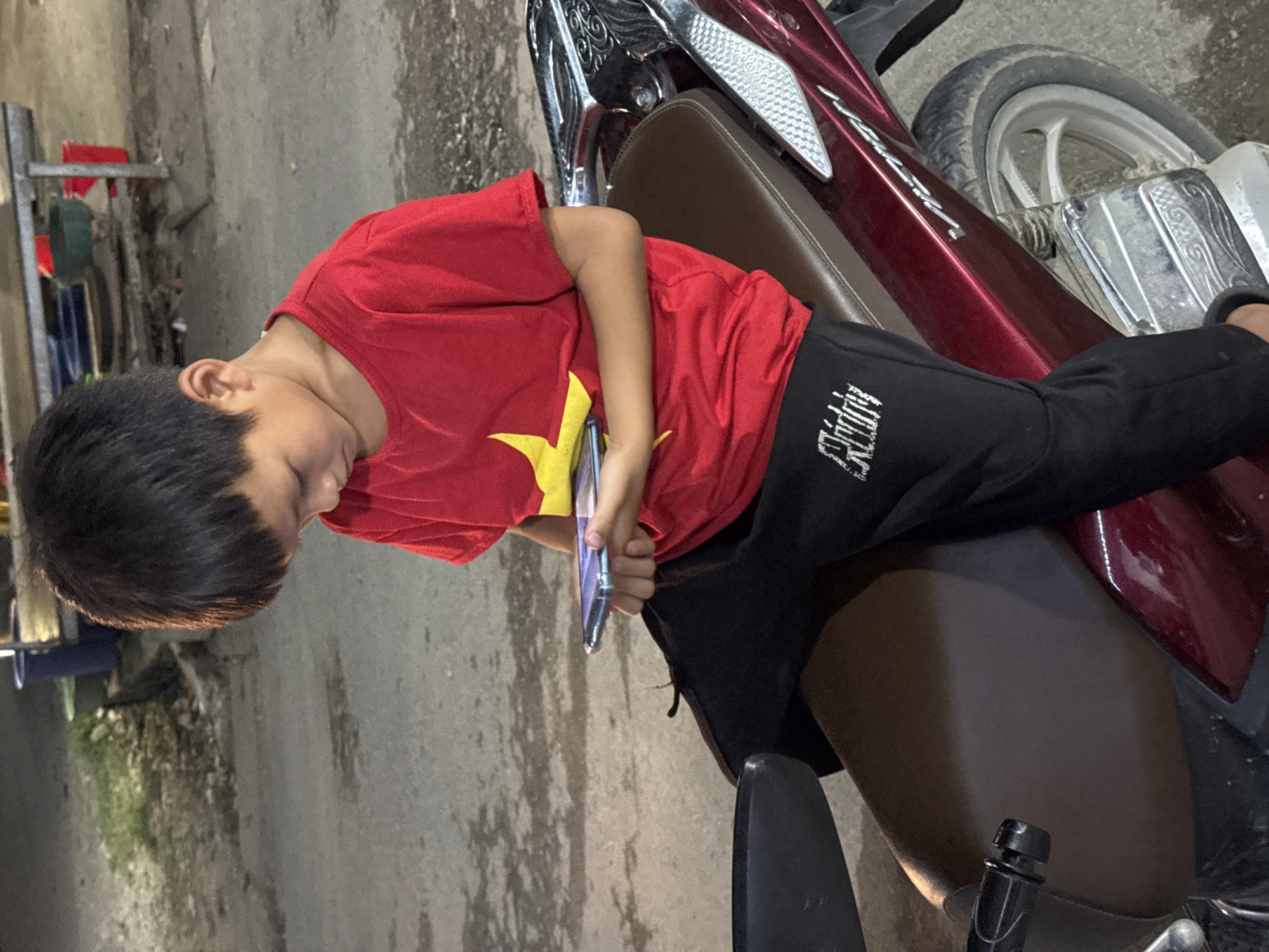 A young boy with short black hair, wearing a red t-shirt with a yellow star and black pants, is sitting on the brown seat of a maroon motorcycle. He is holding a cell phone in his hands. The motorcycle is parked on a paved surface, likely a road. In the background, there is some foliage and what appears to be a metal rack holding various items. The location appears to be Du Già Commune, Vietnam.