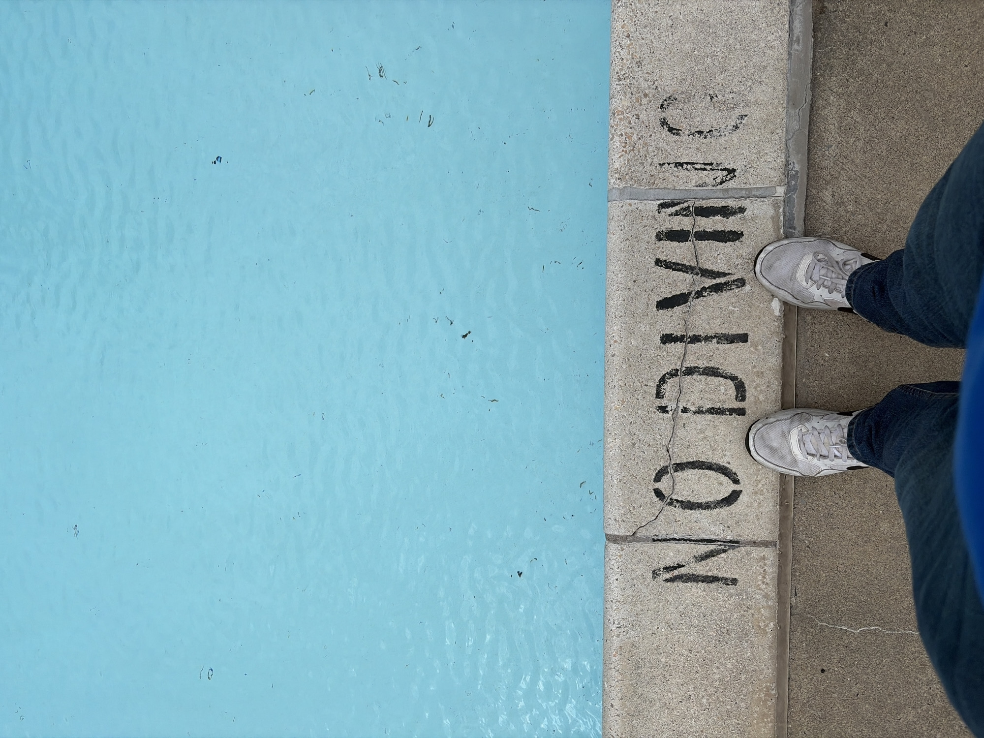 The image shows the edge of a swimming pool with clear blue water. A concrete border with the words "NO DIVING" painted in black is visible. A person wearing blue jeans and grey sneakers stands on the concrete next to the pool. The location of the image is in the city of Edison, United States of America.