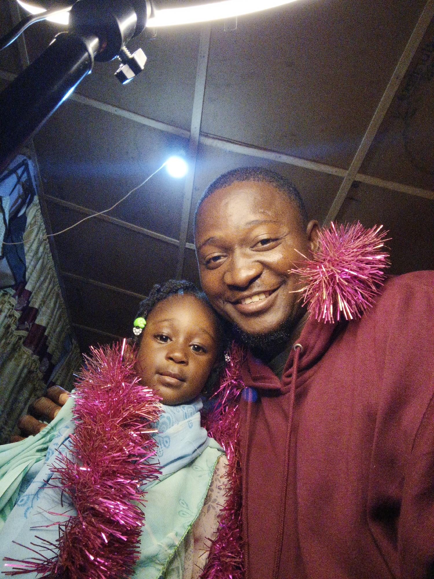 The media file is a close-up, selfie-style photograph of an adult man and a young child. The man is smiling broadly at the camera, and the child is looking slightly off to the side with a neutral expression. Both subjects are adorned with bright pink tinsel, which encircles their necks and is draped over their shoulders. The man is wearing a dark maroon hooded sweatshirt, and the child is wearing a light blue garment, possibly a scarf or shawl, over another patterned fabric.
The background appears to be indoors, with a visible ceiling structure made of corrugated metal or a similar material, and some lighting fixtures. There is also a patterned fabric hanging on the left side of the frame, possibly a curtain or wall covering. The lighting is somewhat harsh, with a bright light source visible in the upper portion of the image, casting some glare. The overall mood seems festive, likely due to the tinsel, suggesting a holiday or celebration. The time of day and weather conditions are not discernible from the image. There is no visible text in the media file.