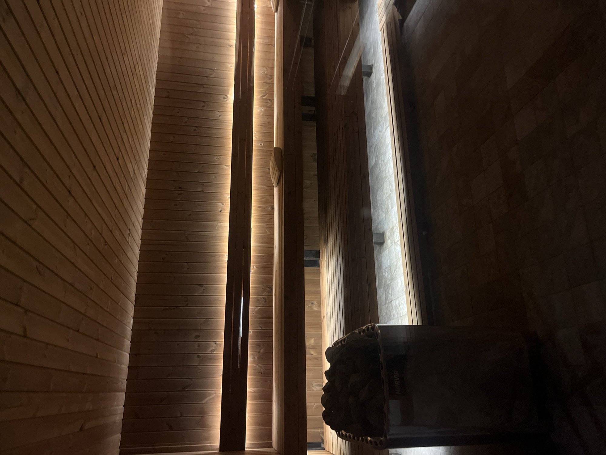 The image shows the interior of a sauna. The walls are lined with light-colored wooden planks, arranged horizontally. Vertical wooden pillars create a sense of depth. Between some of the pillars, there are narrow light sources, possibly LED strips. In the foreground, a dark, rectangular object is visible; it appears to be a Harvia sauna heater, containing dark rocks. The location in Sofia, Bulgaria, is not discernible from the image.