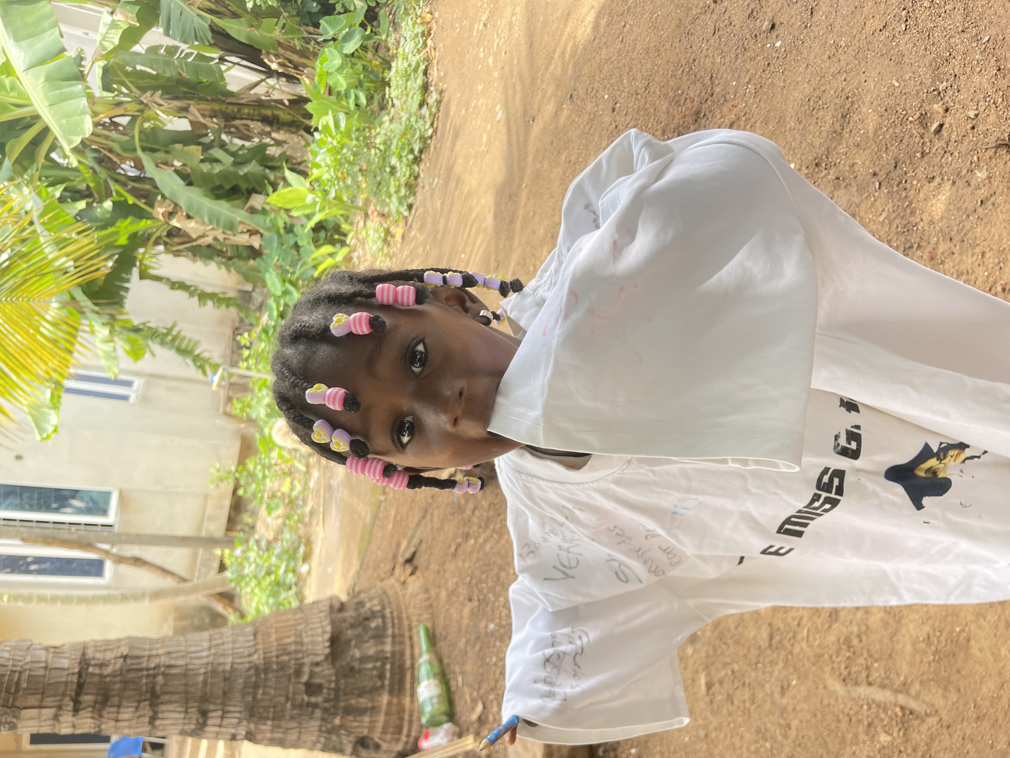 A close-up, upright portrait features a young girl looking directly at the camera. She has dark skin and her black hair is styled in numerous small braids, each adorned with colorful beads in shades of pink, yellow, purple, and black. She is wearing a white t-shirt that bears several handwritten notes and doodles in dark ink; visible text includes "MISS G." and "VEKE," along with other scribbles such as "car" and "on-may." Her right arm is raised, with the white sleeve of her shirt obscuring part of her lower face, suggesting a playful or shy gesture. Her left hand, holding a blue and orange colored pencil or crayon, rests by her side.
The setting is outdoors in what appears to be a sunny, tropical environment, consistent with Akwanga, Nigeria. The ground beneath her is reddish-brown, sandy dirt. In the background, lush green tropical foliage is prominent, including large banana leaves and the textured trunk of a palm tree on the left. A light-colored building with white-framed windows is partially visible behind the vegetation. A green glass bottle can be seen near the base of the palm tree. The bright, clear lighting indicates it is daytime, likely midday or early afternoon. The girl's expression is curious and direct, with a hint of playfulness.