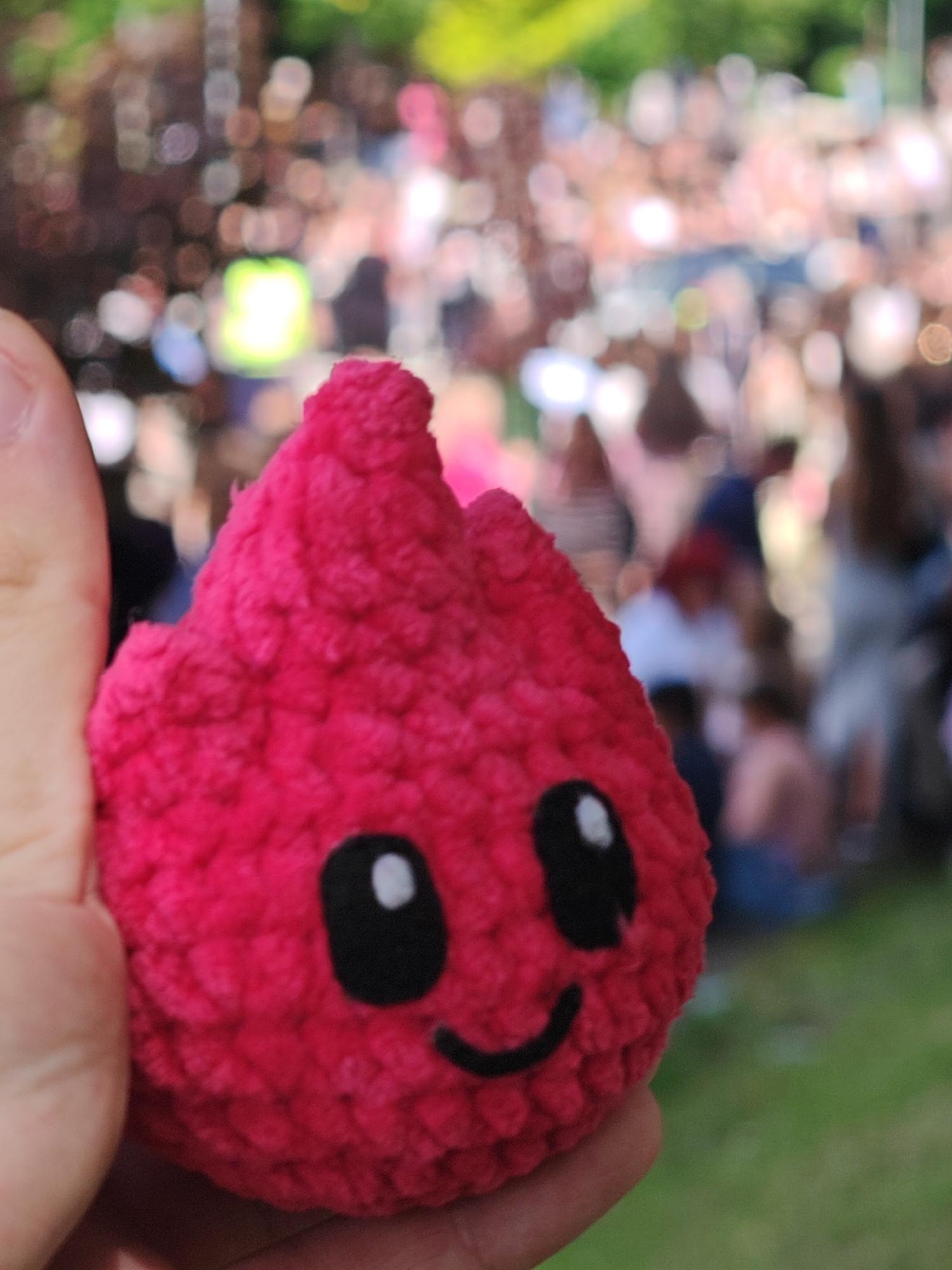 A person is holding a pink, crocheted toy with black eyes and a smiling mouth, resembling a stylized flame. The background shows a blurred scene of a crowd of people, suggesting an outdoor event or gathering, possibly in a park or public space. Green grass is visible in the lower right of the frame. The location in Shumen, Bulgaria, is not definitively indicated in the image.