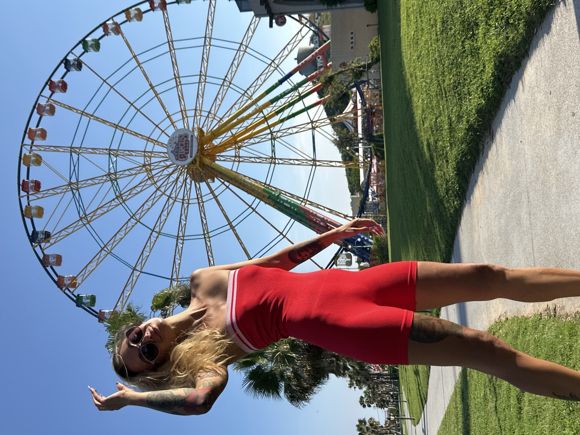 A young Caucasian woman with blonde hair, sunglasses, and a red strapless outfit stands on a paved path in a park. She has tattoos on her arms and legs. Behind her, a large Ferris wheel with colorful carriages is visible against a clear blue sky. Palm trees and manicured grass are also present in the environment. The scene appears to be a sunny day at an amusement park. The location is identified as Tien Thanh Ward, Vietnam.