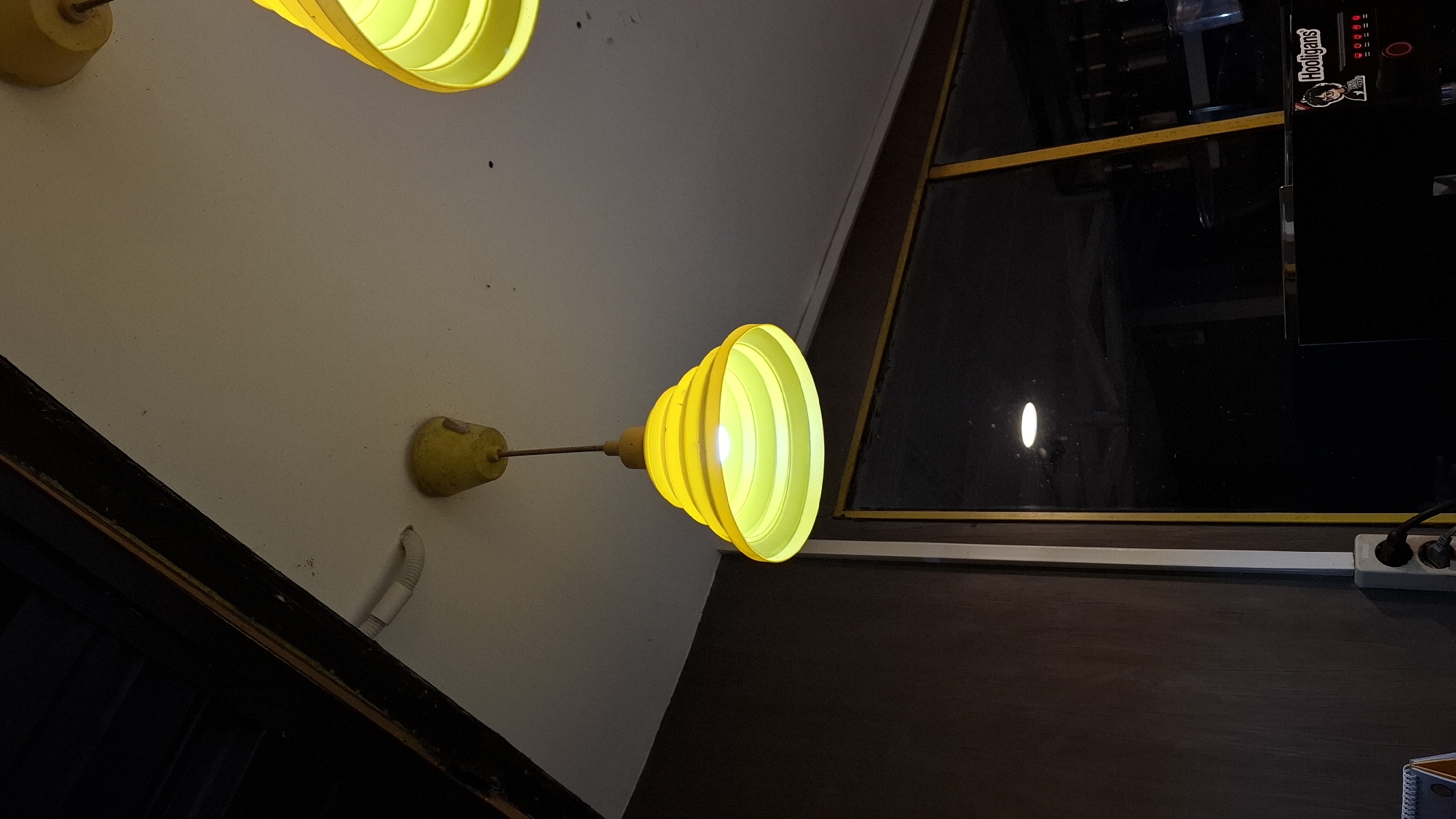 The image depicts an indoor scene, possibly a cafe or restaurant, with a focus on a yellow pendant light fixture. The light fixture has a tiered, conical shade and is suspended from a white ceiling by a thin rod. Another similar yellow light fixture is partially visible in the upper right corner. The ceiling appears to be textured and shows some minor dust or debris.
Below the ceiling, a dark brown wall with a slightly rough texture runs along the left side of the frame. A white electrical conduit runs vertically down this wall, terminating in a double electrical outlet with a plug inserted. To the right of the wall, a large window with a yellow frame is present. The window is dark and reflects some light sources from within the room, including a circular reflection of what appears to be another light fixture.
In the lower portion of the window, a black appliance, possibly a coffee machine, is visible. A sticker with the text "Hooligans" and a cartoon character is affixed to its front. The overall lighting is dim, with the yellow light fixture providing the primary illumination in the central area. The setting is indeterminate in terms of specific location within Unknown, Indonesia, but the presence of electrical outlets and the type of interior decor suggest a commercial establishment.