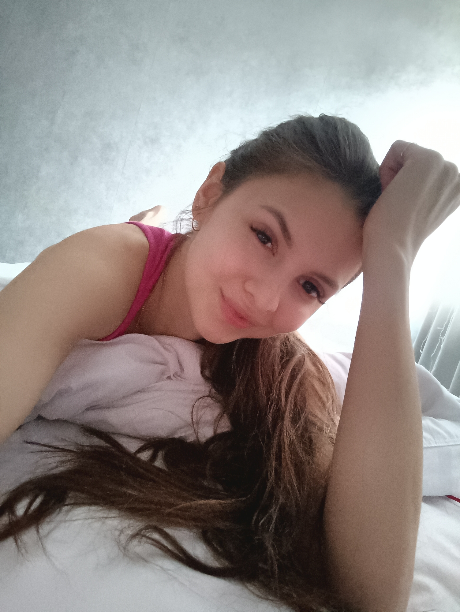 A fair-skinned woman with brown hair pulled back in a partial updo lies on a white bed, resting on a white pillow. She is wearing a pink tank top. Her left arm is extended above her head, with her hand near her forehead. The background is a light grey wall, possibly textured, with a window to the right casting bright light. The location is Chaikovsky, Russia.