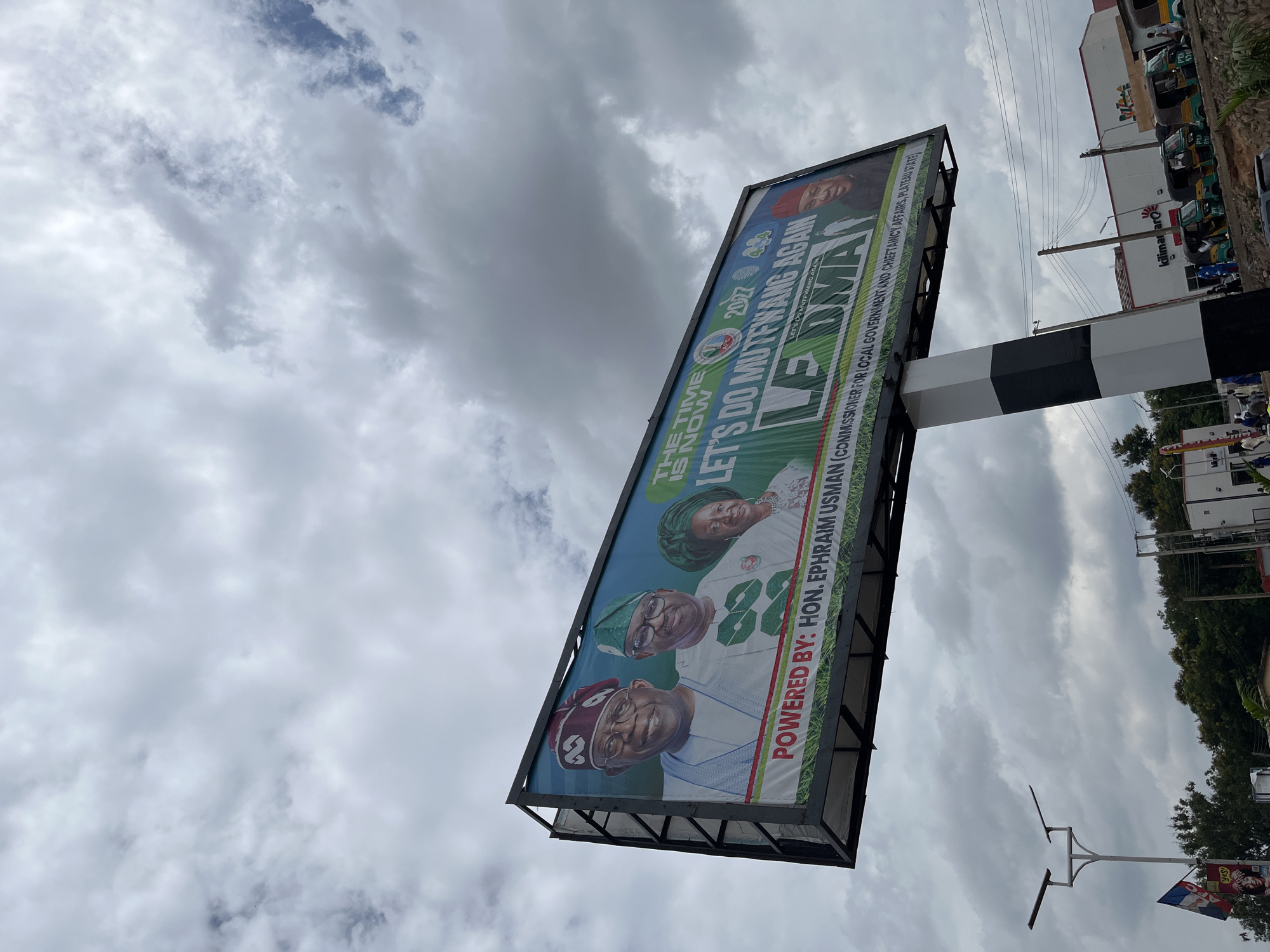 A large billboard stands prominently against an overcast sky in Jos, Plateau State, Nigeria. The billboard's main panel features a light blue background transitioning to green at the bottom. On the left, a smiling man wearing a red-patterned cap and glasses, identifiable as Nigerian President Bola Ahmed Tinubu, is depicted. To his right, another smiling man in a green-patterned cap and glasses, identifiable as Vice President Kashim Shettima, is shown. Adjacent to him is a smiling woman in a green headwrap and attire, identifiable as First Lady Oluremi Tinubu.
The upper right portion of the billboard displays the text "THE TIME IS NOW" in white letters, followed by "2027" and the logo of the All Progressives Congress (APC). Below this, in large white letters, is the slogan "LET'S DO MUTFWANG AGAIN" with "LEO MA" beneath it. On the far right of the billboard, a portrait of a man in a red cap, identifiable as Barr. Caleb Mutfwang, the current Governor of Plateau State, is visible.
At the bottom of the billboard, a white banner with a green grass-like pattern states "POWERED BY: HON. EPHRAIM USMAN (COMMISSIONER FOR LOCAL GOVERNMENT AND CHIEFTAINCY AFFAIRS, PLATEAU STATE)".
The billboard is supported by a tall, black and white striped pole. The sky above is largely covered by dense, light grey clouds. In the background, below the billboard, various urban elements are visible. These include power lines, a street lamp, and several commercial establishments. A building with the "Kilimanjaro" logo and another with the "Pizza Hut" logo are discernible. Multiple green and yellow motor tricycles (keke Napeps) are parked or moving in front of these buildings, typical of Nigerian urban transport. Scattered people and vehicles are present on the street and near the establishments, indicating general city activity. Sparse trees and vegetation are also present in the mid-ground.