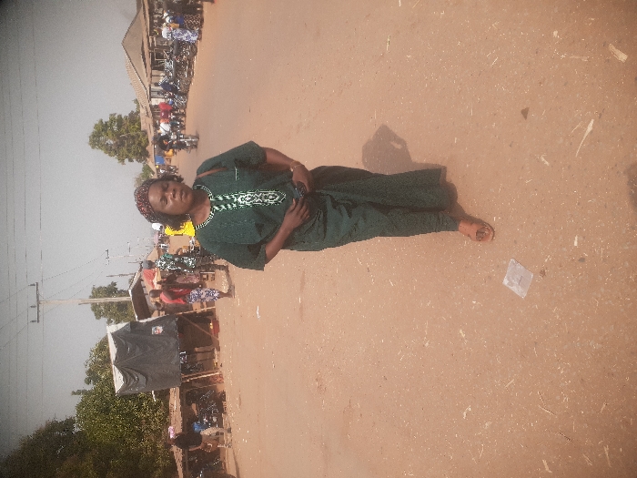 The image displays an outdoor daytime scene on a reddish-brown dirt road in Gwadanye, Nigeria, consistent with a local market or commercial street.
In the foreground, a female individual, likely middle-aged, walks directly towards the camera. She wears a dark green traditional Nigerian tunic with white embroidered patterns on the chest and a matching wrapper. Her head is adorned with a patterned headwrap featuring red, black, and brown hues. Brown, open-toed sandals are on her feet. A dark-colored shoulder bag is visible on her left side, and she holds a dark object, possibly a mobile phone, with both hands clasped in front of her waist. Her shadow extends to her right. The road surface near her feet is uneven laterite, with small white specks and a clear plastic wrapper visible.
The midground and background feature a bustling street environment. To the left, multiple makeshift market stalls or shelters are visible, constructed with wooden frames and covered by dark tarpaulins. Dense green trees and foliage are present behind these structures. A group of motorcycles is parked near the stalls. Several individuals are present, including one wearing a red shirt and another in a camouflage-patterned outfit. Overhead, utility poles with power lines are visible.
To the right side of the road, further simple commercial buildings with corrugated metal roofs line the street. A longer line of parked motorcycles and additional pedestrians are observed on this side, including one individual in a bright yellow top. The road recedes into the distance, appearing active with more people and structures under a bright, slightly hazy sky.