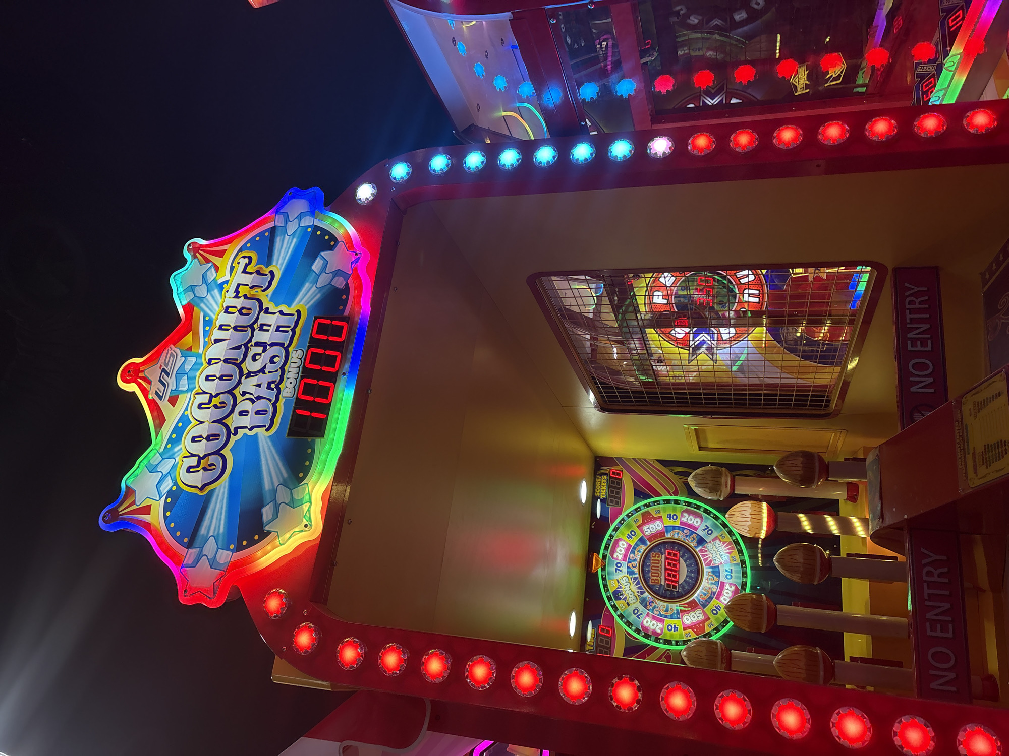 This image captures a vibrant, brightly lit arcade game machine named "COCONUT BASH" in what is contextually Burgas, Bulgaria. The game, likely a redemption or "whack-a-mole" style, features a prominent overhead display illuminating the game's title in colorful neon-like lights, showing "BONUS 1000" in red digital numbers. Below, inside the game enclosure, a circular scoring wheel with segments for points like 40, 70, 200, and 500, along with a "SUPER BONUS" section, is visible. Several brown, coconut-shaped targets on sticks, suggesting a mallet-hitting mechanism, are arranged vertically to the right of the scoring wheel. Digital readouts for "SCORE/ TICKETS" are also present, displaying "8.8.8.8" as a placeholder or current score. The machine is adorned with numerous red and blue LED lights lining its frame, creating a lively and engaging atmosphere typical of an arcade or entertainment center. The background is dark, indicating an indoor setting, possibly an arcade at night or in a dimly lit area. Text like "NO ENTRY" is vertically printed on the side panels. No people are visible in the scene.