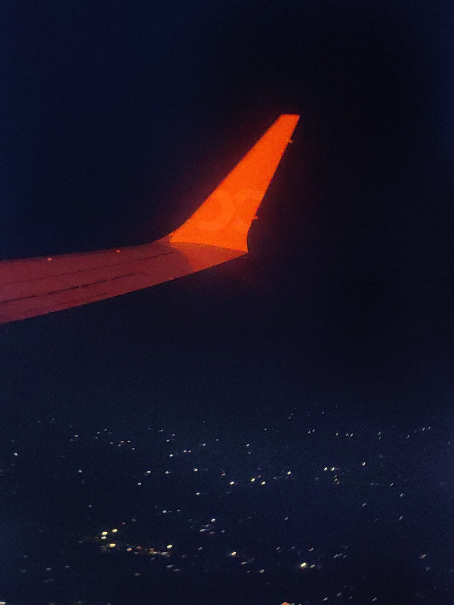 An airplane wing, illuminated by an orange light, extends from the left side of the image against a dark blue night sky. Below the wing, a city is visible with numerous small, scattered lights, indicative of an urban area at night. The lights appear as blurred points of yellow, white, and orange. The scene captures a view from an aircraft at altitude, looking down upon a city, likely Kamruddinpur, India, at night.