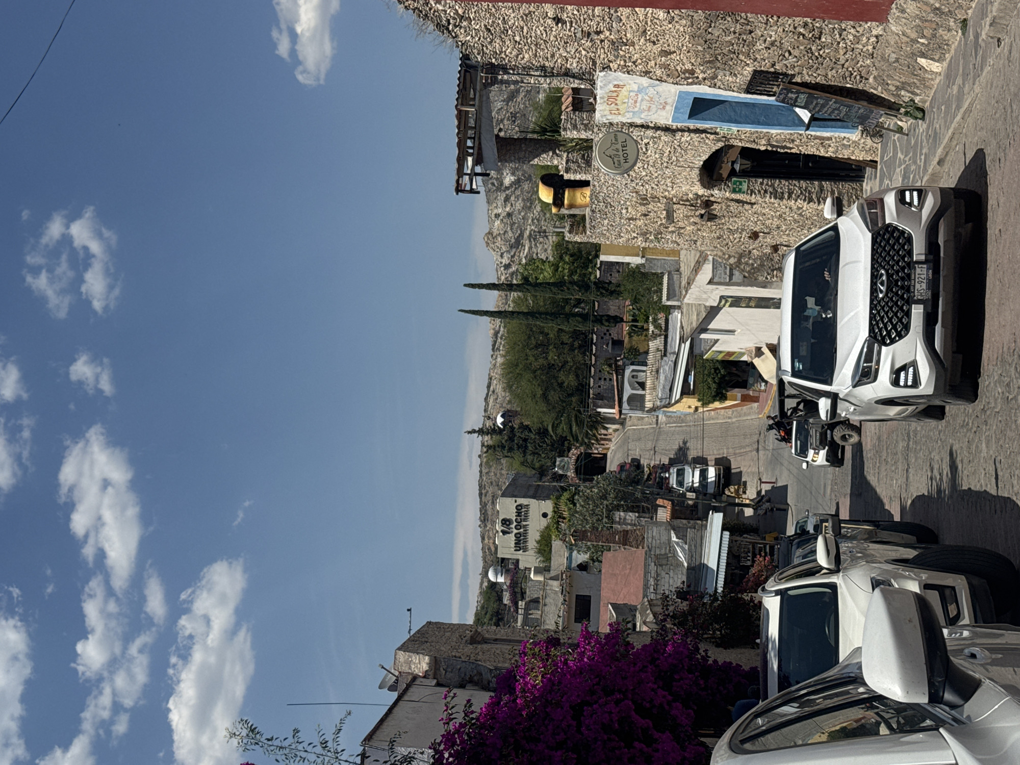 The image presents a daytime urban scene in Bernal, Mexico, characterized by narrow streets and a mix of architecture. The sky is predominantly blue with scattered clouds. The landscape is hilly with buildings constructed from stone and painted in light colors.
In the foreground, there is an abundance of purple flowering bushes alongside white vehicles parked on the roadside, their details clearly visible. Up the street, additional vehicles are parked, including a white off-road vehicle with visible tires.
Signage for "EL SOLAR" and "Casa 19 de Enero HOTEL" is displayed on a stone wall. There's also signage for "1/8 UNO OCHO" on a nearby building.
The photo is taken from an elevated position, with the horizon showing a gradual gradient between the city and the distant mountains. The street curves into the city, where there is a mix of residential and commercial buildings.