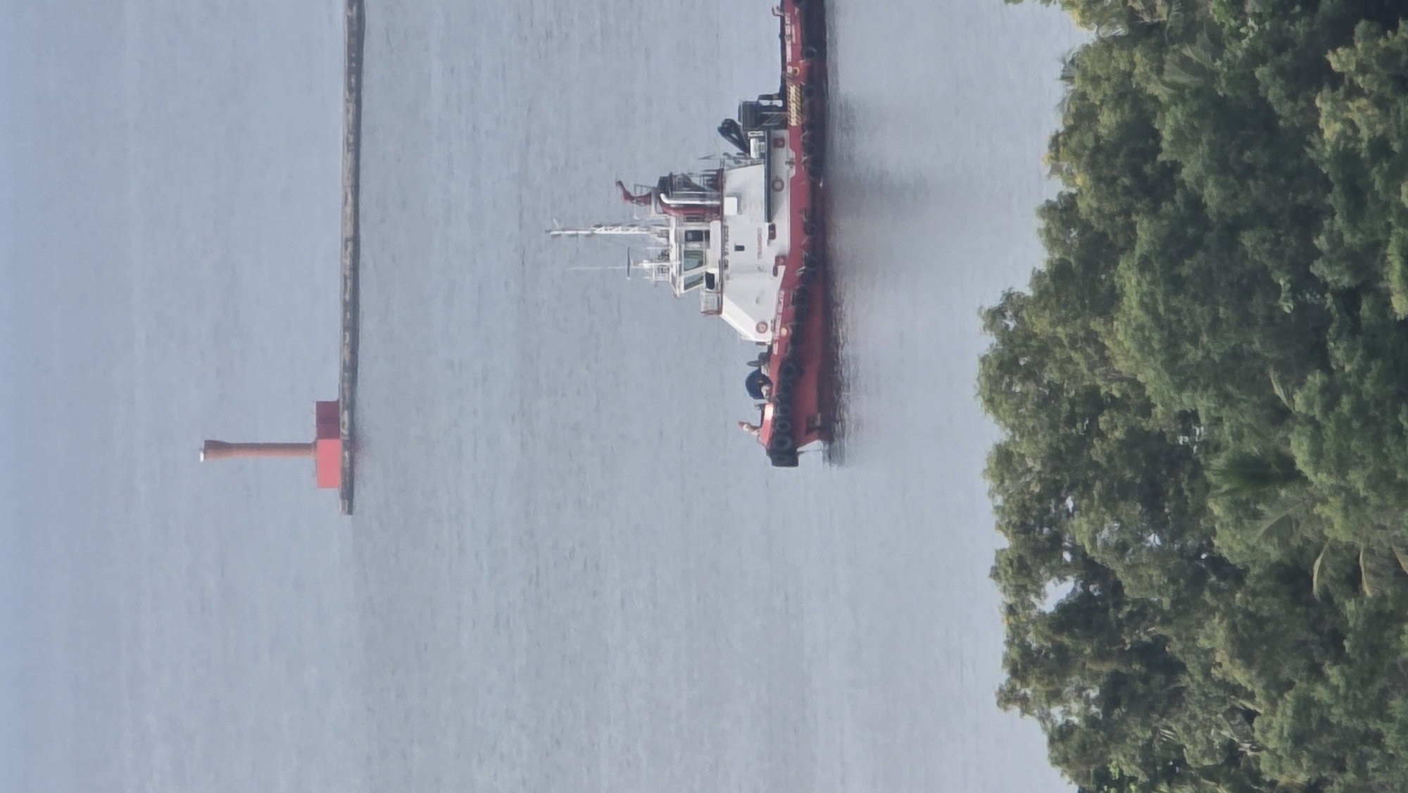 The image shows a tugboat sailing in the waters of Lhokseumawe, Indonesia, next to a pier. The tugboat is red and white. Adjacent to the tugboat is a green, leafy tree. In the distance, there is a gray pier with a red navigational marker.