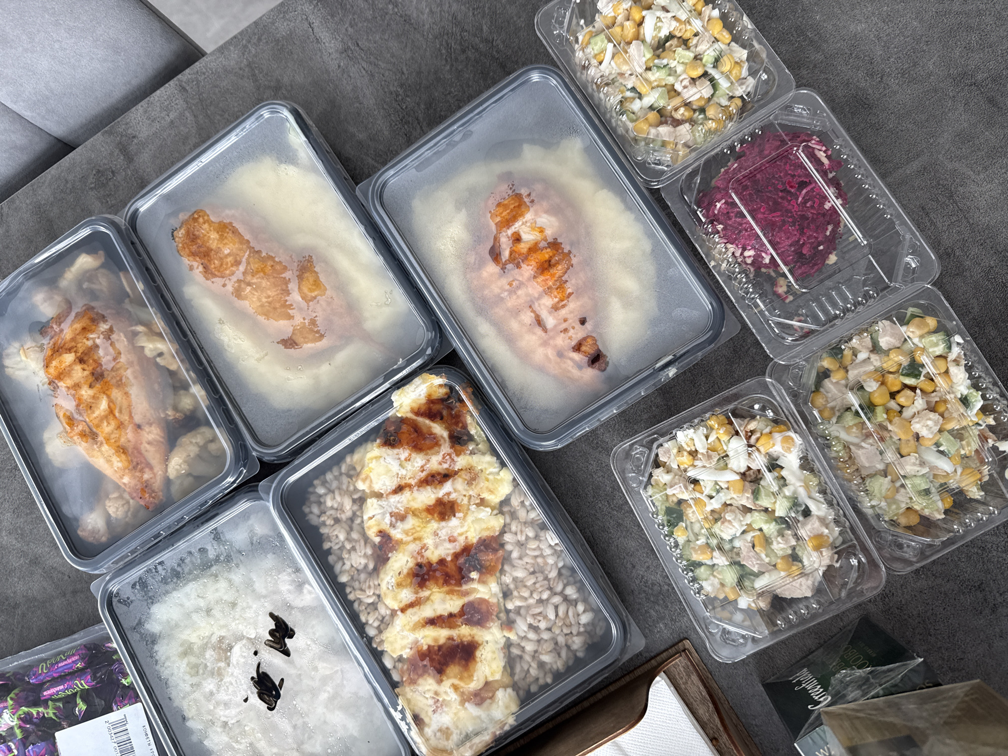 The image captures an overhead view of various pre-packaged meals and other items laid out on a dark gray, textured surface, likely a table or countertop. In the background, a light gray upholstered piece of furniture, possibly a sofa or chair, is visible.
The scene is dominated by multiple transparent plastic containers holding prepared food. There are two larger rectangular containers featuring grilled chicken breasts atop a bed of creamy mashed potatoes, their clear lids showing condensation. Another similar container holds grilled chicken pieces alongside cauliflower florets. A fourth large container features a baked dish with grill marks, possibly an omelet or a grittata, resting on a bed of grains, likely pearl barley. Another rectangular container contains plain white rice with some dark liquid drops on its surface.
Adjacent to these main dishes are three smaller, square-shaped clear plastic containers. Two of them contain a salad composed of corn, diced cucumbers, and what appears to be white meat or chicken, mixed in a light dressing. The third small container holds a vibrant red salad, strongly suggesting beetroot.
To the lower left, a purple plastic bag of candies or chocolates is partially visible. Text on this bag includes "КОНФЕТЫ КРО..." (KONFETY KRO...) and a barcode with the number "2 003247 091017". A green box, partially obscured, is visible near the bottom right, displaying the brand name "GREENFIELD" and part of "WOODB...", suggesting a tea product. A small wooden tray or box with white napkins or tissues is also present in the lower right corner.
The overall impression is of a collection of ready-to-eat meals, suggesting meal preparation or a food delivery service, consistent with a setting in Krasnoyarsk, Russia.