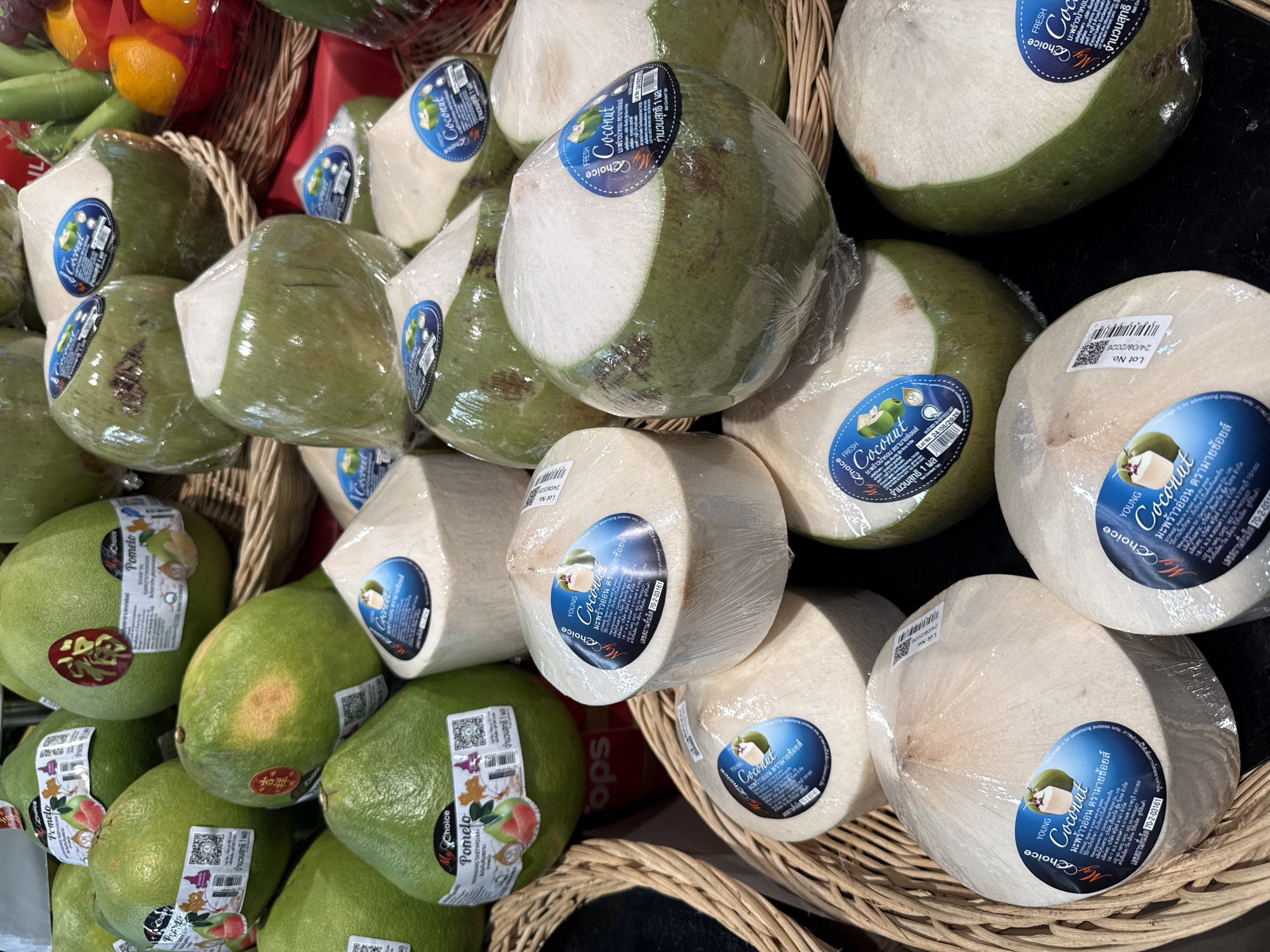 The image shows a close-up, high-angle view of a fruit stand in a market, possibly in Pattaya City, Thailand, displaying a variety of fresh fruits. The foreground is dominated by numerous young coconuts, many of which are wrapped in clear plastic. These coconuts are stacked and arranged in wicker baskets. Several of the coconuts have blue, oval-shaped labels that read "Young Coconut" and include Thai text and a lot number. In the background, several large green pomelos are visible, also with labels and some appear to be wrapped. Some of the pomelos have red stickers with Chinese characters. The overall impression is one of abundance and freshness, typical of a tropical fruit market. There are no people visible in the image. The lighting suggests an indoor or shaded outdoor setting, possibly a market stall.