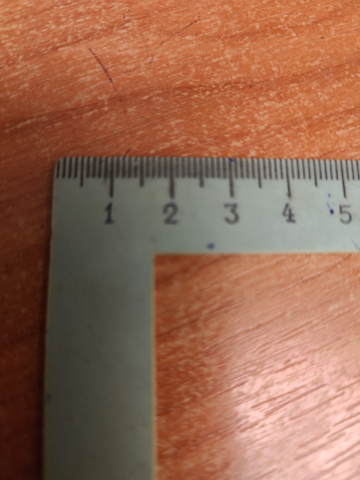 A close-up, overhead shot shows a light blue ruler with black markings placed on an orange, wood-grain surface. The ruler has millimeter and centimeter markings. The numbers 1, 2, 3, 4, and 5 are clearly visible, aligned with centimeter increments. There are also small blue marks scattered on the ruler and the surface. The surface appears to be a table or desk. The image is slightly blurry, particularly the background.
