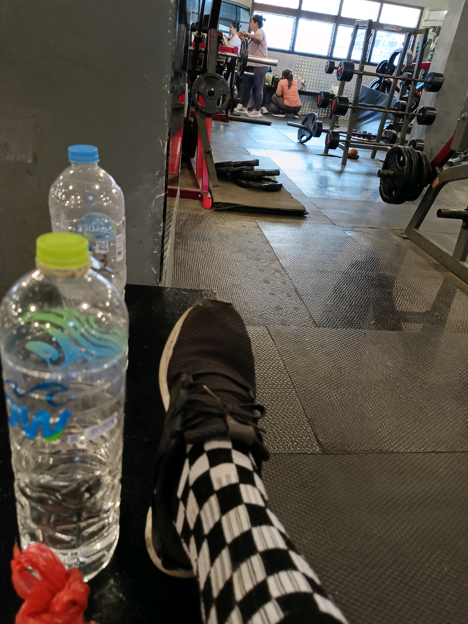In an indoor gym, the foreground features a person's legs, clad in black and white checkered socks and black athletic shoes, resting on a black mat. To the left, two clear plastic water bottles stand upright, one with a blue cap and the other with a green cap, both filled with water. A red object, possibly a gym accessory, lies next to the bottles. The background reveals a gym environment with weightlifting equipment, including barbells, weight plates, and racks. Several individuals are visible in the background, engaged in exercise or moving around the gym. The floor is covered with dark, textured mats. The lighting suggests an indoor setting with natural light filtering in from windows at the far end of the gym.