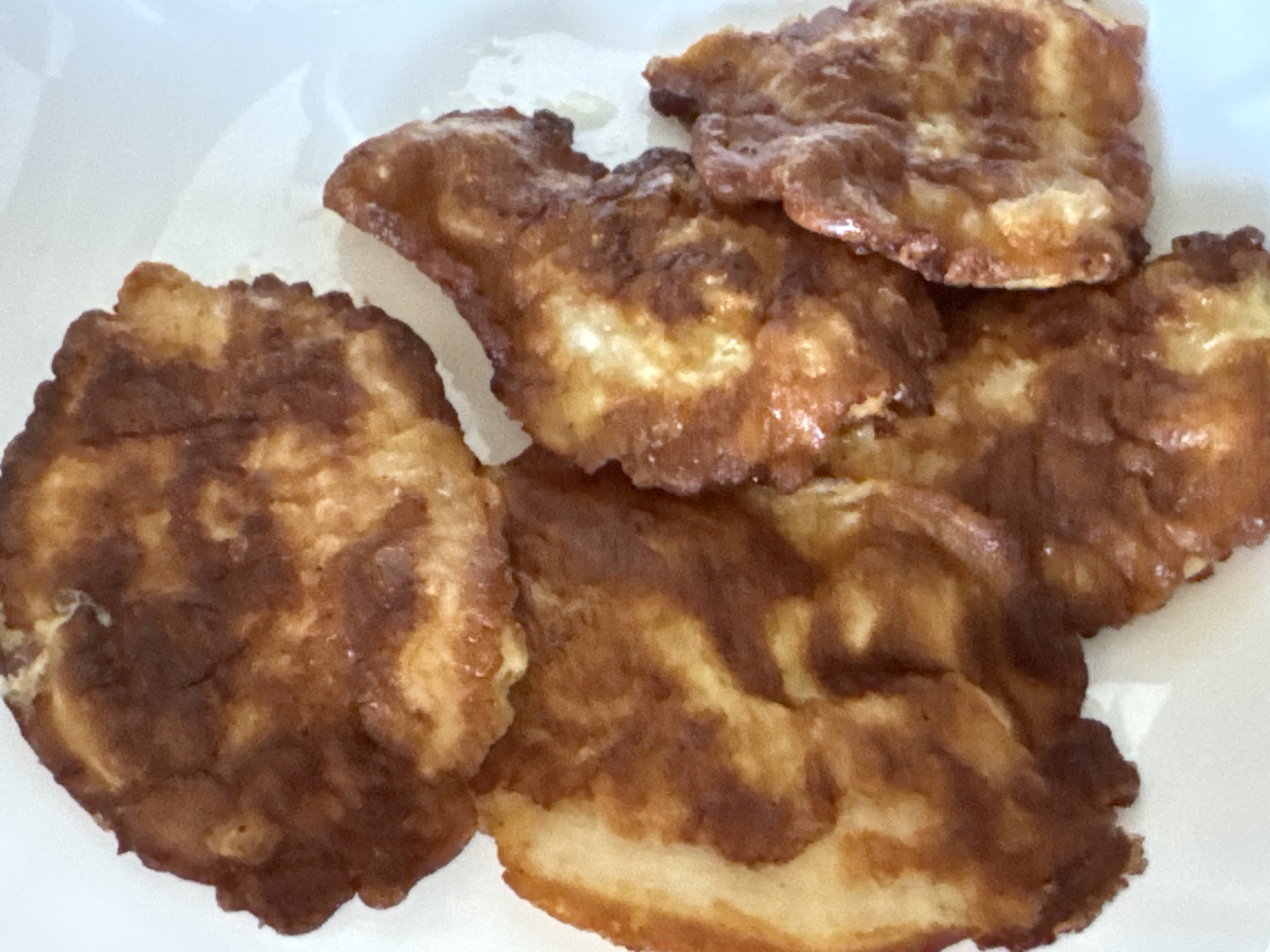 A close-up, overhead shot showcases a serving of golden-brown, fried chicken cutlets on a white plate. The cutlets vary in size and shape, with irregular edges and a slightly uneven texture, indicating they were likely pan-fried. The frying process has created patches of darker brown and lighter golden hues across the surface of the chicken, giving it a rustic appearance. Some areas show a slightly glossy sheen, suggesting oil from the cooking process. The cutlets are layered and overlapping, with the bottom cutlet appearing the largest and most intact. The white plate provides a simple backdrop, allowing the rich color of the chicken to be the main focus. No people, text, or specific location cues are visible. The lighting is even, suggesting an indoor setting, possibly a kitchen or dining area.