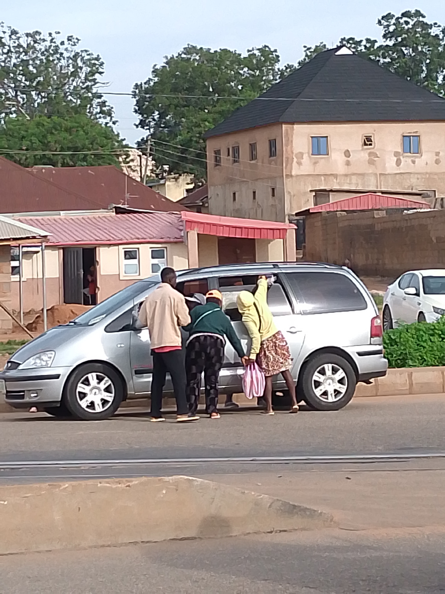 In Kuru Dungus, Nigeria, three individuals are engaged near a silver minivan on a paved road. Two figures, one wearing a light brown jacket and red shirt, and another in a green jacket and patterned pants, stand beside the open driver's side door. A third person, in a yellow hooded jacket and a patterned skirt, reaches into the minivan from the rear passenger side. The minivan has its rear passenger door and sliding door open. In the background, buildings with reddish-brown roofs and a larger building with a dark, multi-faceted roof are visible. Green trees line the background, and a white sedan is parked to the right of the minivan. The scene appears to be a street in a developed area.