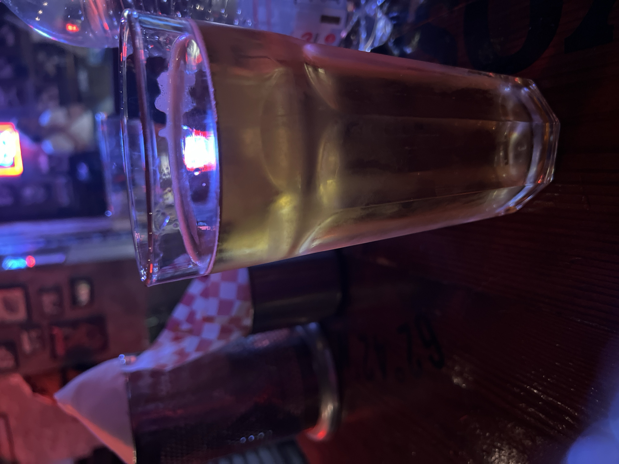 A glass of beer is on a dark wooden surface, likely a bar top. A red and white checkered napkin is visible near the base of the glass. Reflections and ambient lighting suggest an indoor environment, possibly a bar or restaurant. There are dim lights and blurred images on the periphery. A partial number "6.2...29" is etched or printed on the wood near the right of the glass. The location may be Tbilisi, Georgia, although no definitive sign exists to confirm this.