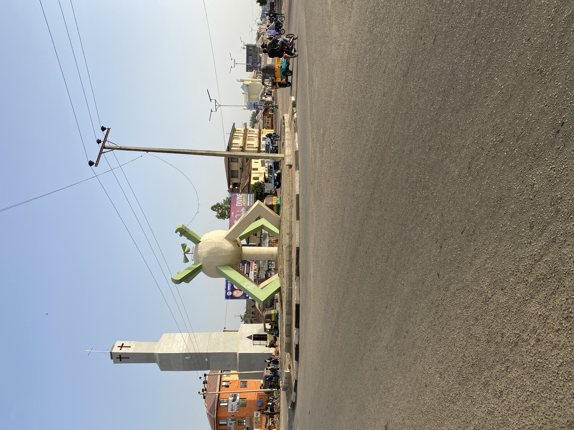 The image shows a street scene in Jos, Nigeria. In the foreground is a sculpture of a round object with green, arm-like extensions. Behind it is a tall, modern church tower with crosses on its facade. A utility pole stands nearby with power lines stretching across the sky. The street itself is asphalt, and there are buildings lining the sides. Motorbikes and auto rickshaws are visible along the road and on the side of the road. People are present on the sidewalks, although their details are difficult to discern. The sky is clear and blue.