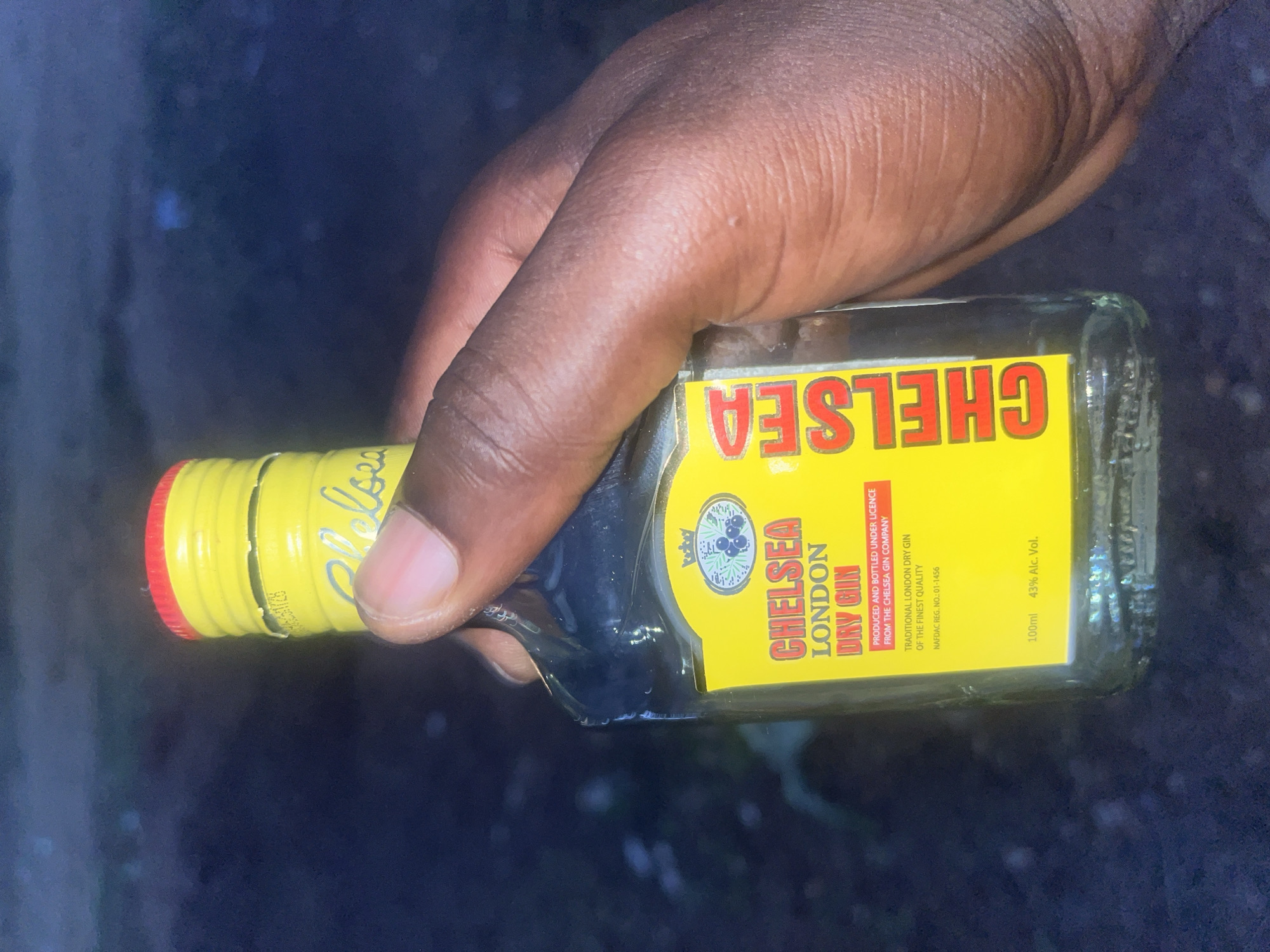 A person's dark-skinned hand is holding a small bottle of Chelsea London Dry Gin. The bottle has a yellow label with the word "CHELSEA" in red capital letters. The label also indicates that the gin is produced and bottled under license from The Chelsea Gin Company. The bottle has a yellow and red cap. The background is blurred and dark, but appears to be an outdoor setting. Based on information on the label indicating "NAFDAC REG. NO.: 01-1456," which is a registration number for the National Agency for Food and Drug Administration and Control in Nigeria, the image is likely taken in Nigeria, potentially Offatedo.