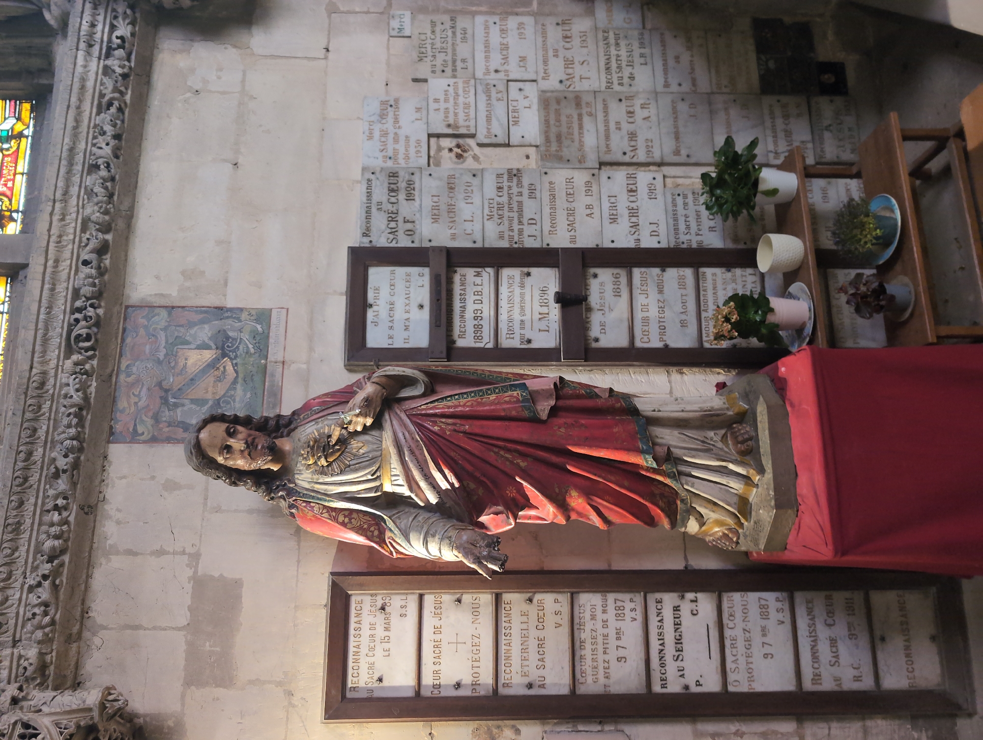 The image depicts an interior view of a church, likely the Église Saint-Rémi in Dieppe, France, based on similar imagery. Dominating the left side of the frame is a full-body statue of Jesus Christ, rendered in polychrome sculpture. Jesus is shown with his right hand outstretched, palm open, and his left hand holding a Sacred Heart from which flames emanate. He is adorned in a red robe with gold trim over a white tunic and appears to be standing on a red cloth-draped pedestal.
To the left of the statue is a tall, narrow wooden frame containing multiple engraved plaques, each likely a votive offering or prayer. These plaques have French text, such as "RECONNAISSANCE AU SACRE CŒUR DE JÉSUS" (Gratitude to the Sacred Heart of Jesus).
The background wall is made of light-colored stone blocks. A portion of a stained-glass window is visible at the top of the frame, and above the plaques, a framed painting hangs on the wall, depicting a coat of arms.
To the right of the statue, a cluster of similar engraved plaques, also in French and likely votive, are mounted directly on the wall or in lighter-colored frames. Some of these plaques are dated, with inscriptions like "1920," "1919," and "1940."
Below the plaques and to the right of the statue, a wooden shelf unit holds several potted plants, including a pink and green plant in a pink pot, a white ceramic pot, and a white pot with a green plant. Another shelf below holds two smaller potted plants. The overall impression is one of a sacred space with devotional elements and greenery.