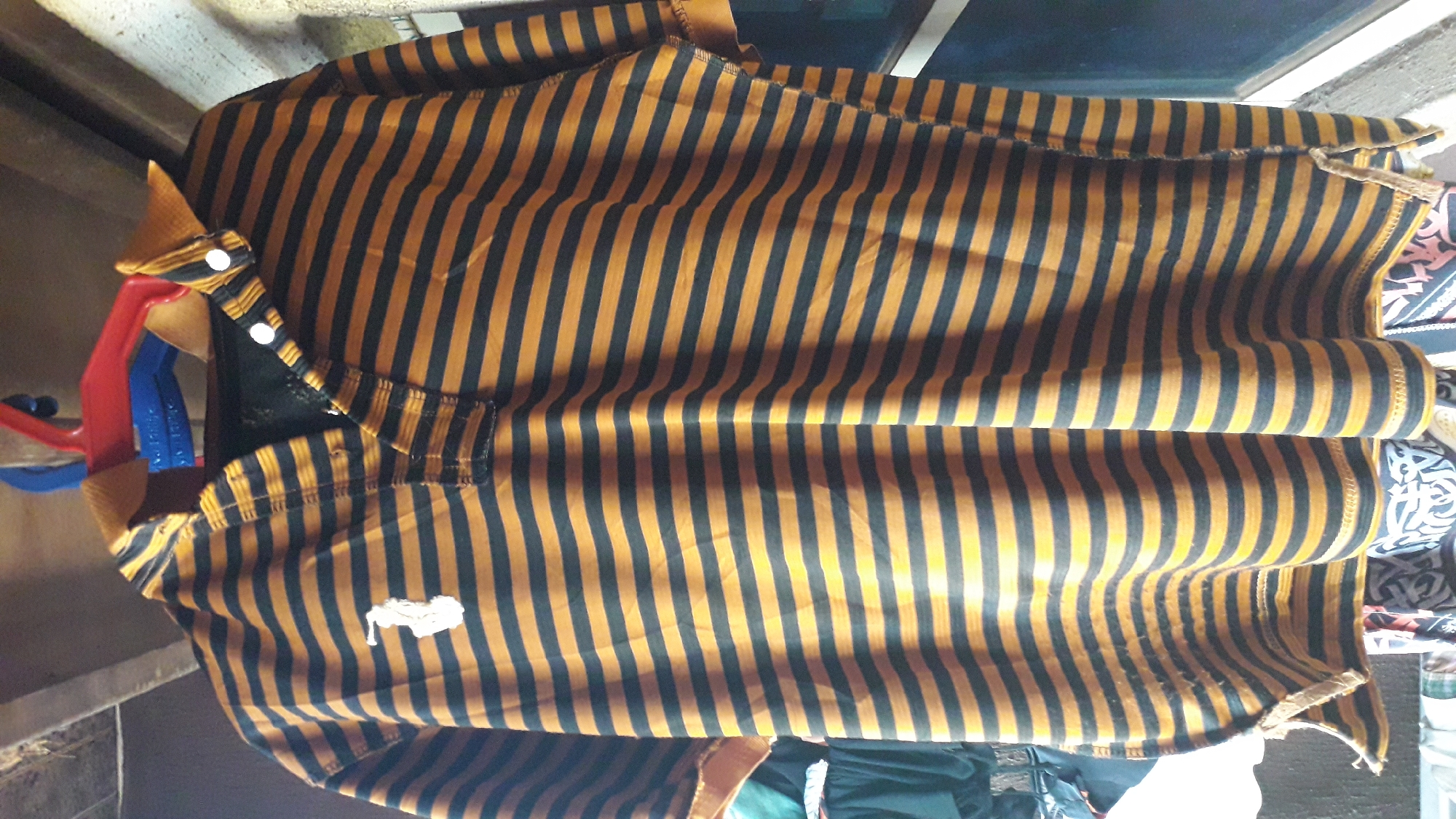 The image shows a striped polo shirt with black and orange stripes. The shirt has a white logo on the lower left front. It's hanging with white buttons visible on the collar. The shirt is being held by a red and blue plastic clamp. The location is in Nkpor, Nigeria.