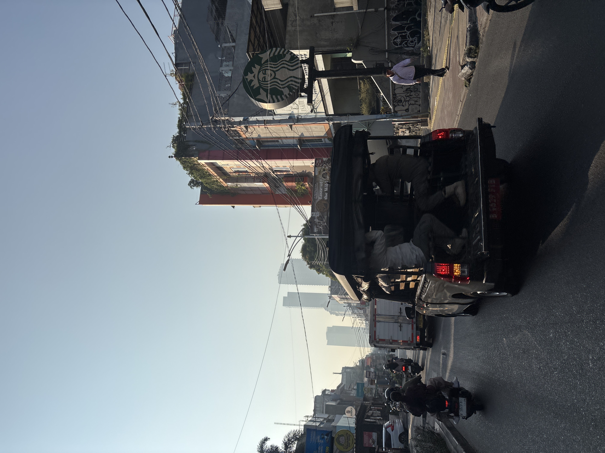The image depicts a street scene in West Jakarta, Indonesia, characterized by moderate traffic under a clear sky. In the foreground, a black pickup truck with a canopy is seen driving on the road, with several individuals seated in the back. To the left, a motorcycle with a rider and passenger moves along the same road. Further ahead, more motorcycles and a white truck are visible, contributing to the traffic flow. On the right side of the street, a building features a prominent Starbucks sign. A man in a light-colored jacket and dark pants walks on the sidewalk next to the Starbucks. The surrounding environment includes various buildings, power lines, and some vegetation. The overall atmosphere suggests a typical urban street during daytime.