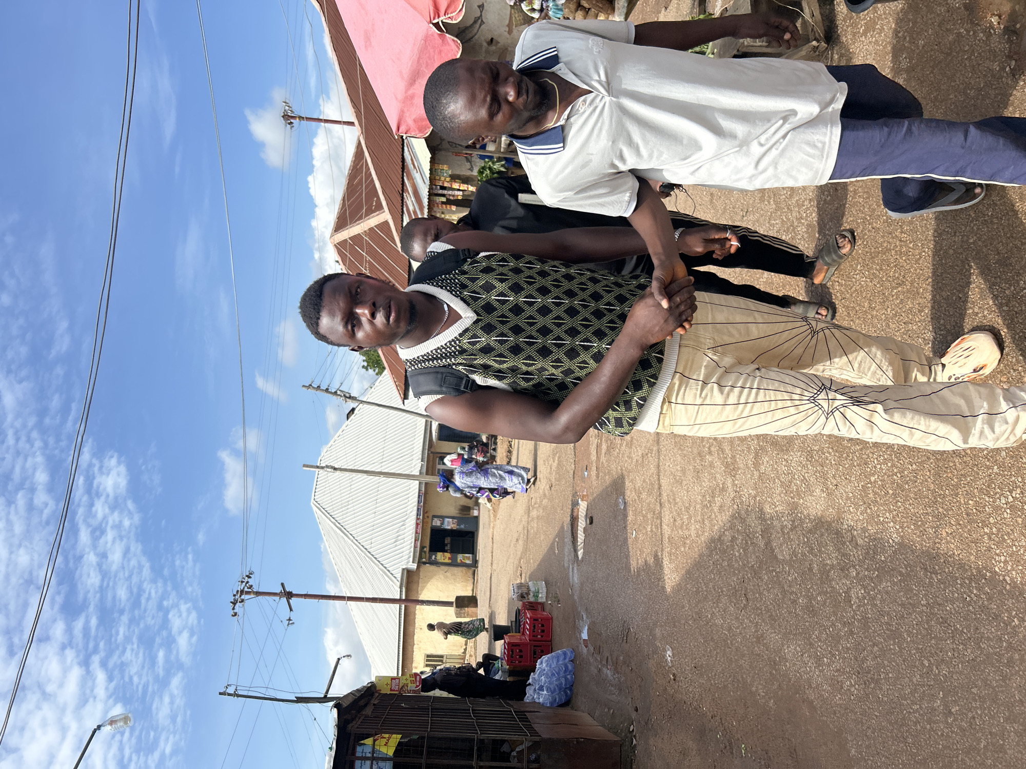 In Jos, Nigeria, under a bright, cloudy sky, a group of men are engaged in an interaction on a street. Two men are prominently featured in the foreground, with their hands clasped as if in a handshake or greeting. One man is wearing a white polo shirt with a blue collar and trim, and dark blue track pants. The other man is dressed in a black and green patterned vest over a white shirt, and light-colored pants with a geometric design.
Behind them, other individuals are visible, and in the background, there are buildings, market stalls, and a pink umbrella. Red crates and bottles of water are on the ground, suggesting a commercial or market setting. Power lines crisscross the sky, indicating an urban or semi-urban environment. The lighting suggests it is daytime. The overall scene appears to be a casual street encounter or transaction. No visible text is discernible.