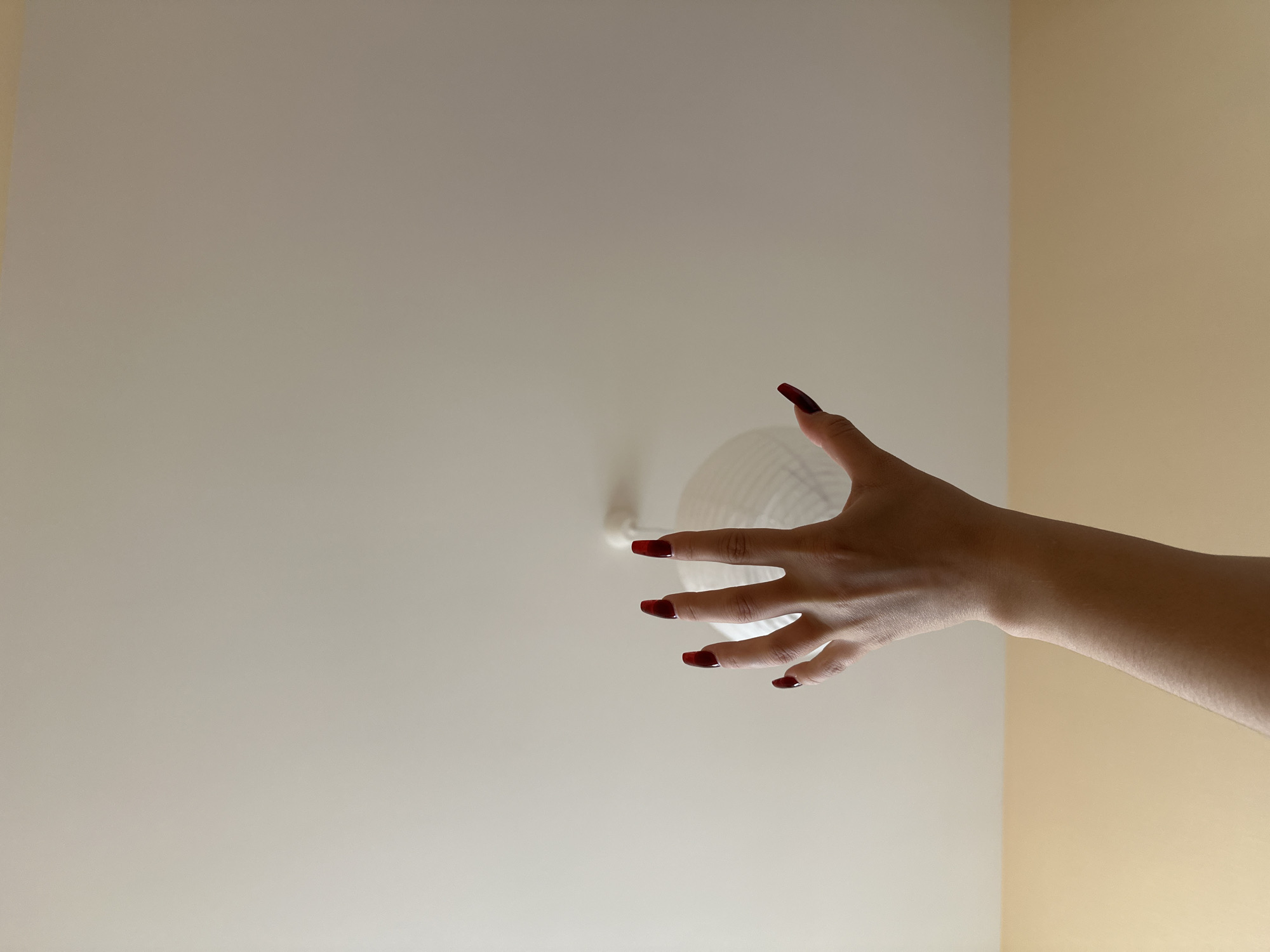 An arm and hand reach toward a ceiling light fixture. The hand has red nail polish. The wall color changes from pale beige to a darker beige. The location and city are unknown.