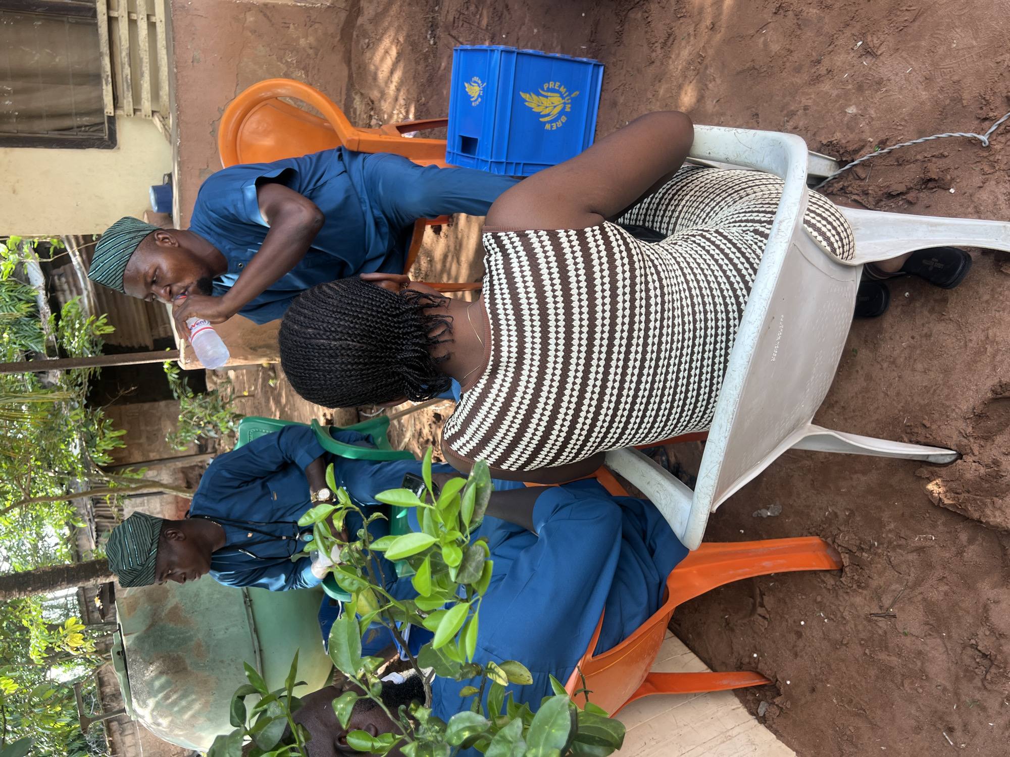 Location: Nkpor, Nigeria.
Scene: An outdoor gathering with several individuals seated on plastic chairs.
People: Two men are dressed in matching blue uniforms and green caps. One man is drinking from a plastic bottle. A woman is wearing a vertically striped brown and white dress.
Objects: Plastic chairs in orange, green, and white. A blue plastic crate with a yellow logo. A water tank. A small tree or bush.
Actions: People are sitting and socializing.
Environment: Outdoor area with bare ground. The scene appears to be in a residential or semi-public space. A building with a window.
