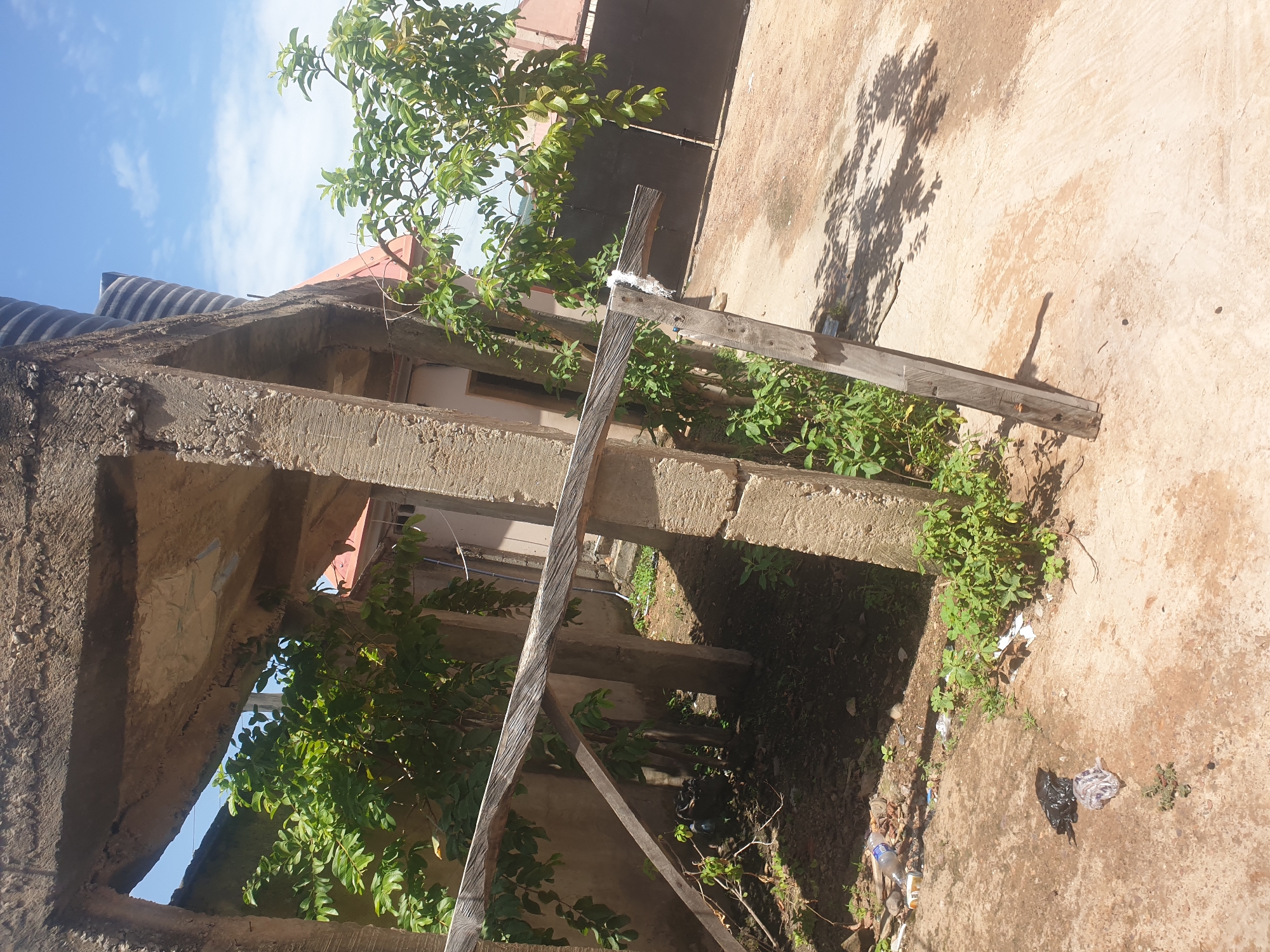 The image shows an overgrown, unfinished concrete structure with a partially collapsed roof in Lokoja, Nigeria. A makeshift wooden support system, possibly intended for scaffolding, leans against the concrete pillars. Vegetation is abundant, encroaching on the structure and surrounding ground. Litter is scattered on the ground, indicating neglect or abandonment. The sky is visible in the background. There are no people present in the image.