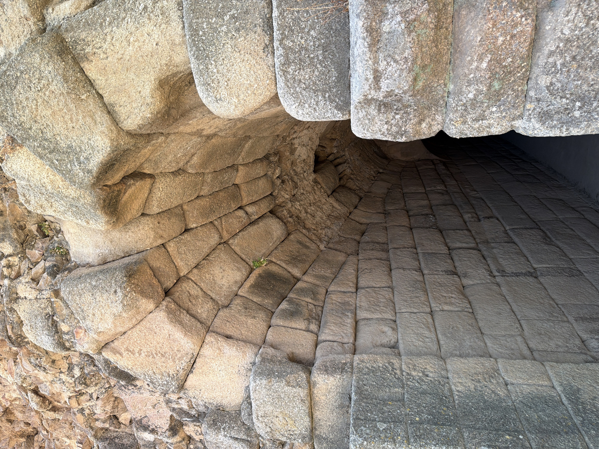 This is a close-up, eye-level, medium shot of the arched entrance to an underground passage within the Roman amphitheater in Merida, Spain. The passage walls are constructed from rough-hewn, irregular stone blocks, creating a textured and aged appearance. Sunlight illuminates the left side of the entrance, casting shadows into the dark, narrow interior of the passage. The archway itself is formed by larger, more precisely cut stone voussoirs, some of which are partially obscured by the rougher stonework of the surrounding structure. The scene is static and devoid of people or animals. The overall impression is one of ancient architecture and a sense of depth extending into darkness.