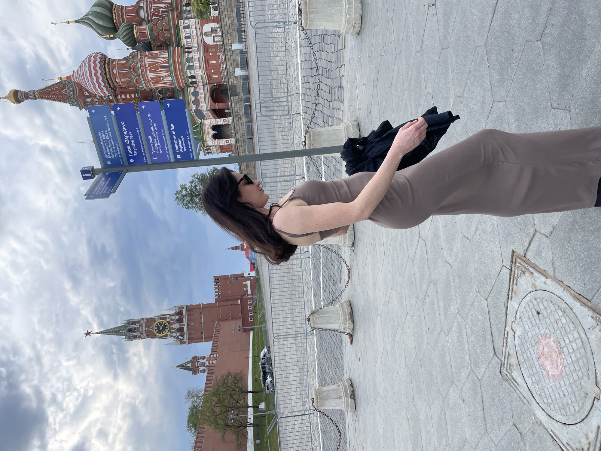 The image shows a woman in a brown dress standing near Red Square in Moscow, Russia. In the background are St. Basil's Cathedral and the Kremlin towers. There is a sign with Cyrillic writing pointing toward the Church of Maximus the Blessed, Zaryadye Park, a Tourist Information Center, and Zaryadye Hall. The woman is holding a black garment. The foreground shows a paved pedestrian area with fences and a manhole cover.