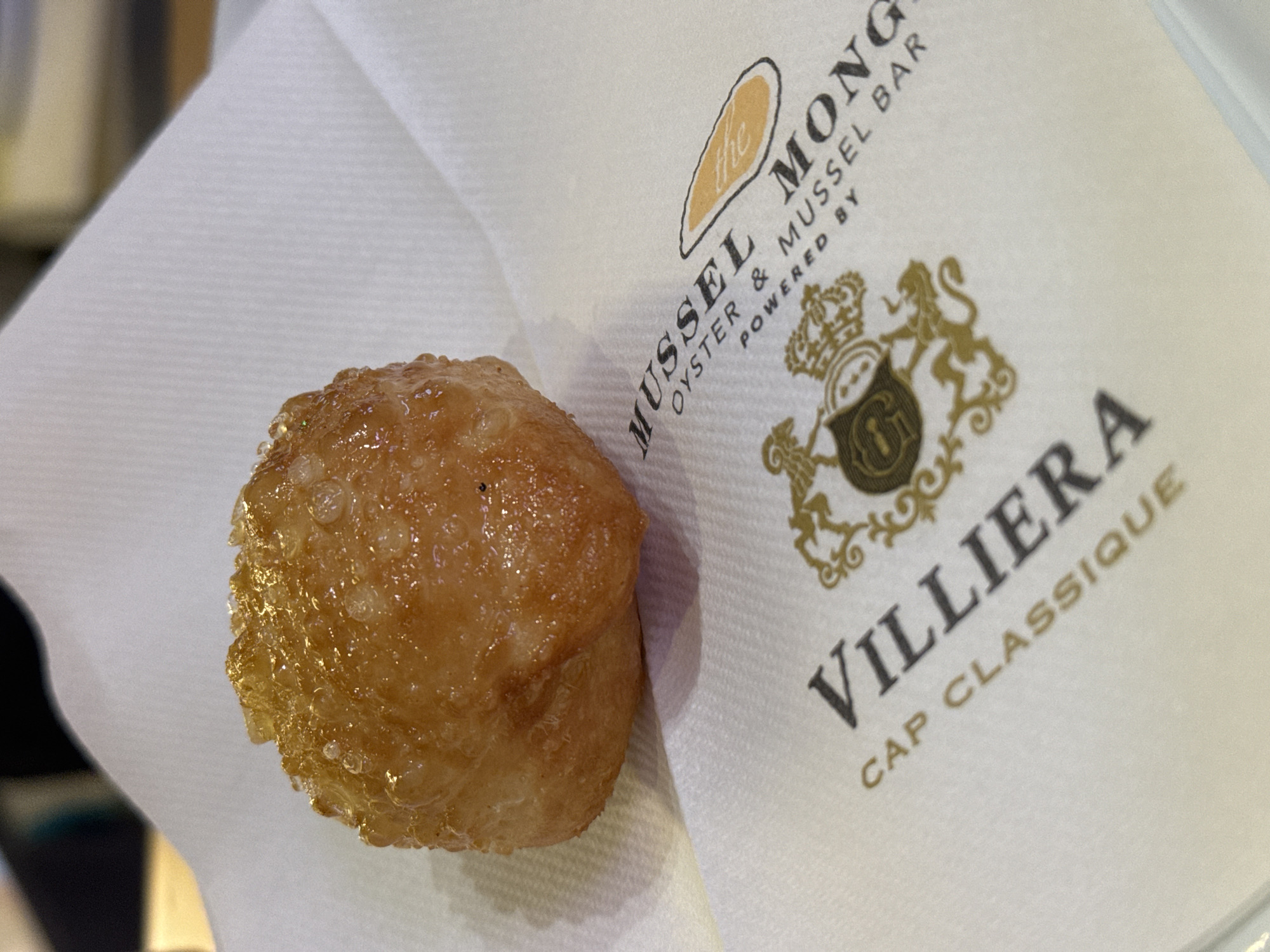 The image shows a glazed donut on a white napkin with the logo of "The Mussel Monger Oyster & Mussel Bar powered by" and the brand "Villiera Cap Classique". This combination indicates that the location of the scene is likely in Cape Town, South Africa, given that Villiera is a wine estate in the Cape Winelands. The primary object is the donut, which appears golden and sugary. The napkin suggests the donut was served at the establishment indicated by the logo. The background is slightly blurred, indicating a shallow depth of field.