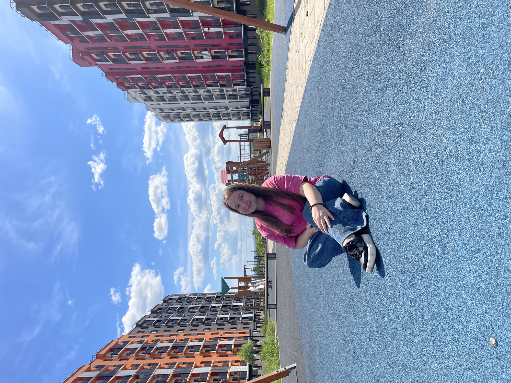 The image depicts an outdoor scene in Pervomaysky, Russia, under bright daylight. A young adult female, with long dark brown hair, is seated cross-legged on a textured, light blue rubberized playground surface, positioned centrally in the lower half of the frame. She wears a short-sleeved pink t-shirt with small red patterns, blue denim jeans, and black sneakers with white soles. A black beaded bracelet is visible on her left wrist. She is looking directly at the camera with a slight smile.
In the background, two multi-story residential apartment buildings flank a playground area. The building on the left features orange, dark gray, and white facades with numerous balconies. The building on the right exhibits magenta, dark gray, and white facades, also with multiple balconies. Between these structures, various wooden and metal playground equipment, including slides and climbing frames, are visible. A long wooden bench is situated in the mid-ground. A light-colored paved pathway runs diagonally across the mid-ground, separating the blue surface from grassy areas with scattered green foliage. The upper portion of the image is dominated by a bright blue sky with numerous scattered white cumulus clouds.