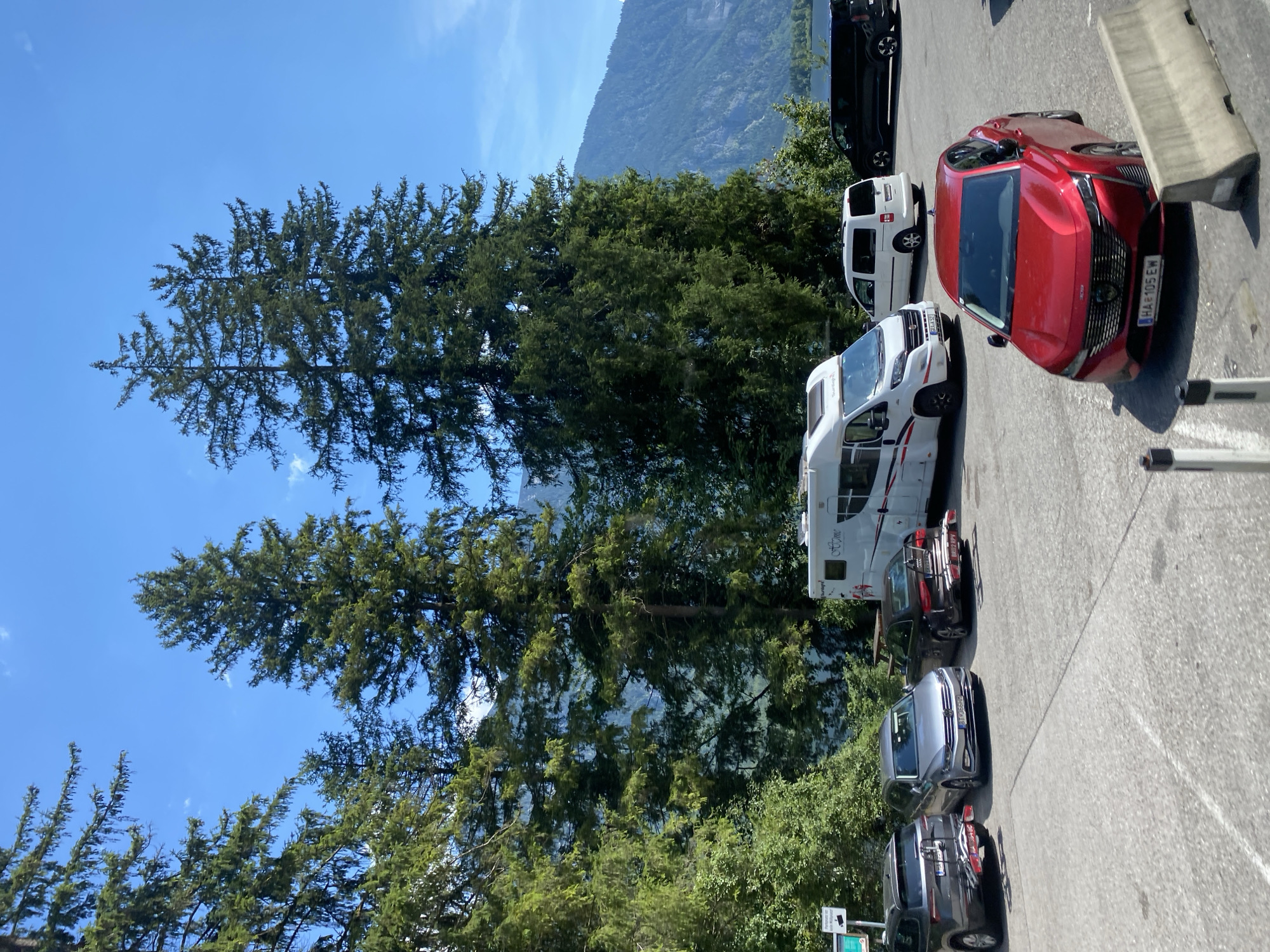 A parking lot is situated in front of a scenic view of a lake and mountains. Several vehicles are parked in the lot, including a white motorhome, a red sedan, a grey station wagon, a dark SUV, and a black van. Tall pine trees are scattered throughout the scene, casting shadows on the ground. The sky is blue with a few scattered clouds. A sign is visible on the left side of the image, indicating parking information. The location is identified as Bad Goisern am Hallstättersee, Austria.