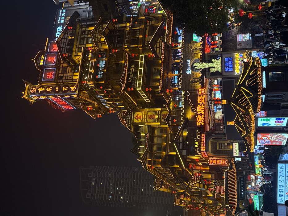 The image displays the Hongyadong architectural complex at night, located in Yuzhong District, Chongqing, China. The complex comprises multiple tiers of buildings built into a hillside, exhibiting traditional Chinese architectural styles with tiled roofs, prominent eaves, and timber elements.
The structures are extensively illuminated by artificial lighting. Warm yellow LED strip lights outline the architectural features and rooflines, while numerous red lanterns hang from eaves and balconies. Multiple red and blue neon signs displaying Chinese characters are affixed to the building facades, with a particularly tall, pagoda-like tower section prominently lit at the top of the central building. A modern, dark high-rise building with illuminated windows is visible in the upper left background, contrasting with the traditional architecture.
At the lower right and foreground, a significant crowd of pedestrians is present, occupying a brightly lit public area. Street-level kiosks featuring illuminated advertising boards, some displaying images of food (e.g., fruit), are visible. A sign on the lower left indicates "洪崖洞景区停车库" (Hongyadong Scenic Area Parking Garage). A glowing green decorative sculpture is situated above a stone archway at a lower entrance level. Trees with visible foliage are present on the far right.