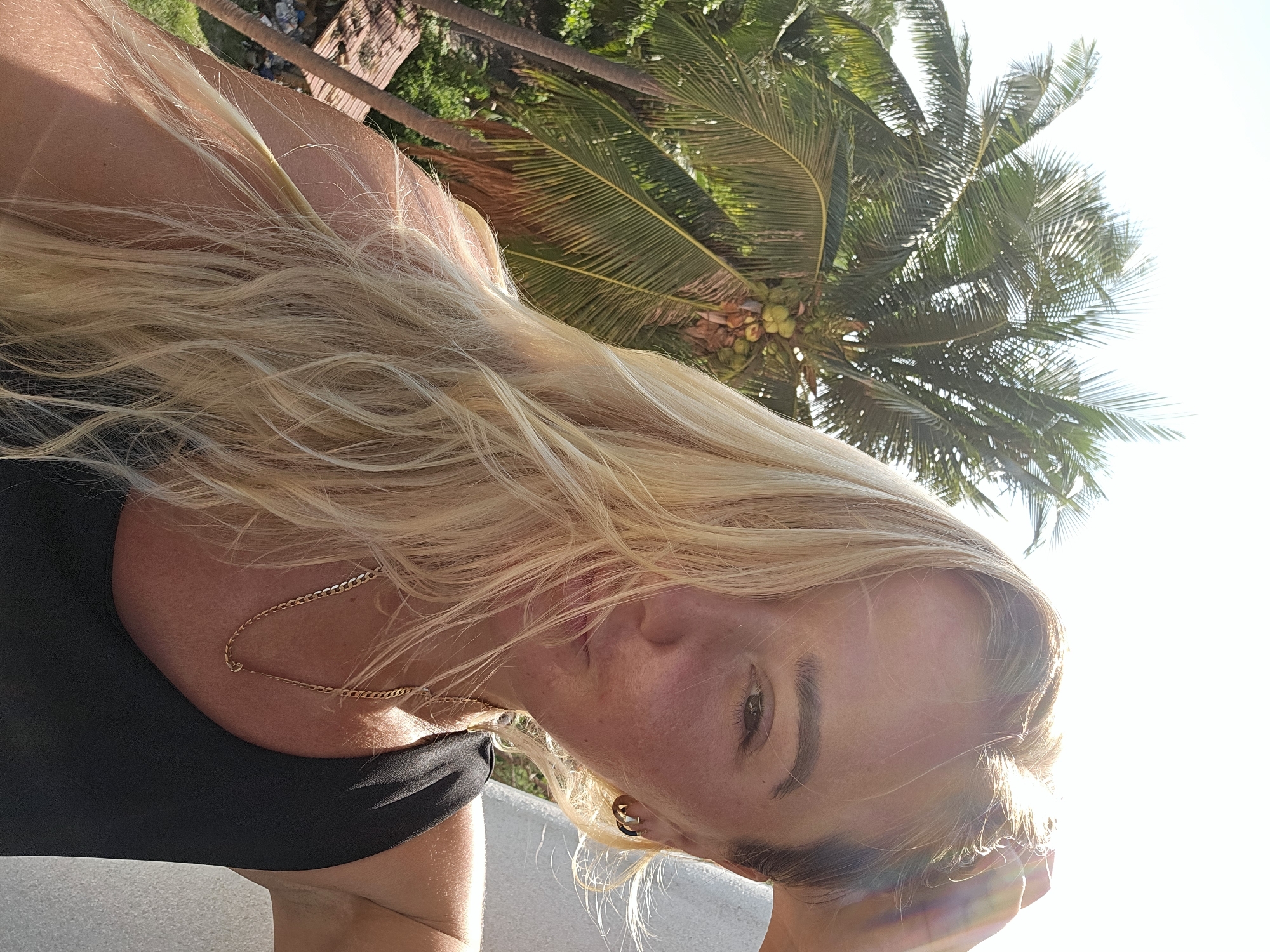 A woman with blonde hair is taking a selfie outdoors with palm trees in the background. She is wearing a black top and a gold chain necklace. The sun is bright, casting a glare on her face and hair. She is touching her hair with her right hand. The location is not specified as Phu Thuy Ward, Vietnam.