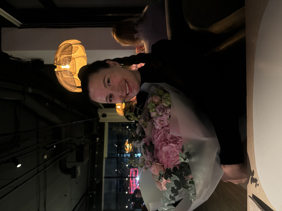 A woman smiles, holding a bouquet of pink and white flowers. She's seated at a table inside what appears to be a restaurant. Her dark hair is styled in braids. Soft lighting from a decorative lamp illuminates the scene. A partially visible person is seated behind her. The restaurant's interior features visible pipes and a window with city lights.