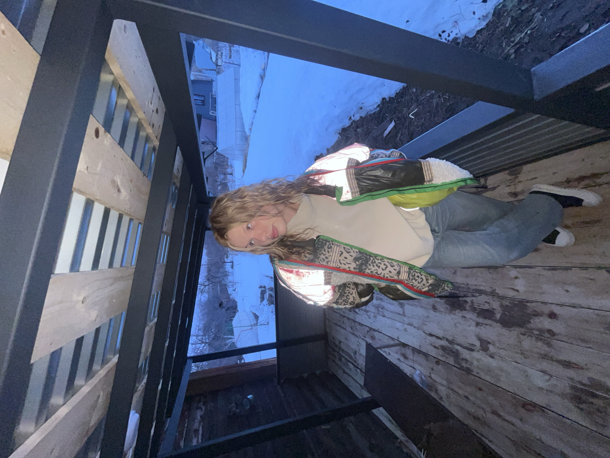 A young person with long blonde hair is lying on a wooden surface within a structure, likely a balcony or a partially enclosed space. The person is wearing jeans, a light-colored sweatshirt, and a patterned jacket with a white and green lining. The structure consists of black metal framing with wooden planking. Outside the structure, there is a view of a landscape with snow, buildings, and trees, suggesting an urban or suburban setting. Based on the context provided, this scene is potentially located in Chaikovsky, Russia.