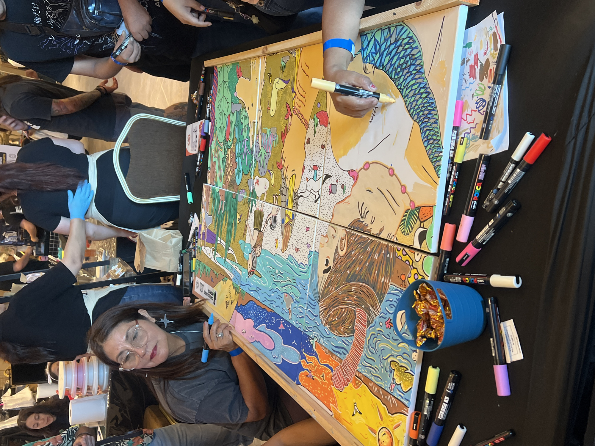The image captures a scene of collaborative art creation in Arica, Chile. Two individuals are actively working on a large, horizontally oriented artwork composed of two connected canvases. The artwork features a vibrant, illustrative style with depictions of nature and fantastical creatures. One person, wearing a blue wristband, is using a yellow marker on the upper canvas, while another person is using a blue marker on the lower canvas, both working on separate sections of the joint art piece.
Several other individuals are present, observing and interacting with the art creation process. One woman with glasses is positioned near the lower canvas, appearing to be engaged in the process. Various markers of different colors are scattered around the artwork on a black surface. A blue cup filled with candy is also visible on the table. The surrounding environment appears to be an indoor space with other people and items, suggesting a public setting like an art event or workshop.