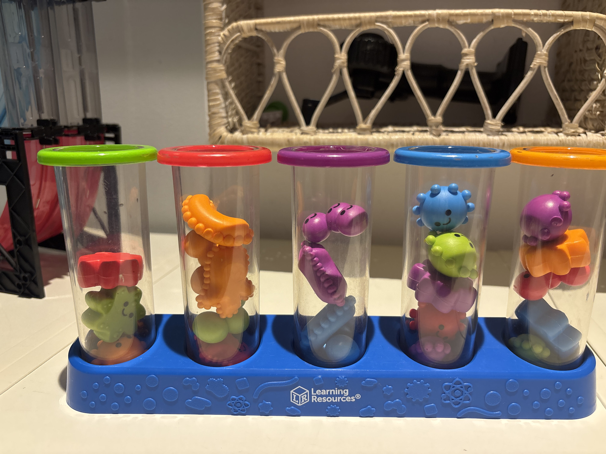 The image shows a close-up, eye-level, indoor shot of a Learning Resources "Stacker" toy. The toy consists of a blue base with circular indentations, holding five clear plastic tubes with colored lids: green, red, purple, blue, and orange. Each tube contains a stack of brightly colored, textured plastic shapes, resembling baby toys. The background is out of focus and appears to be a domestic setting with a woven basket shelf visible. There are no people present in the image. The lighting is soft and diffused. The image is not geotagged and provides no information about its location in Dubai, United Arab Emirates.
